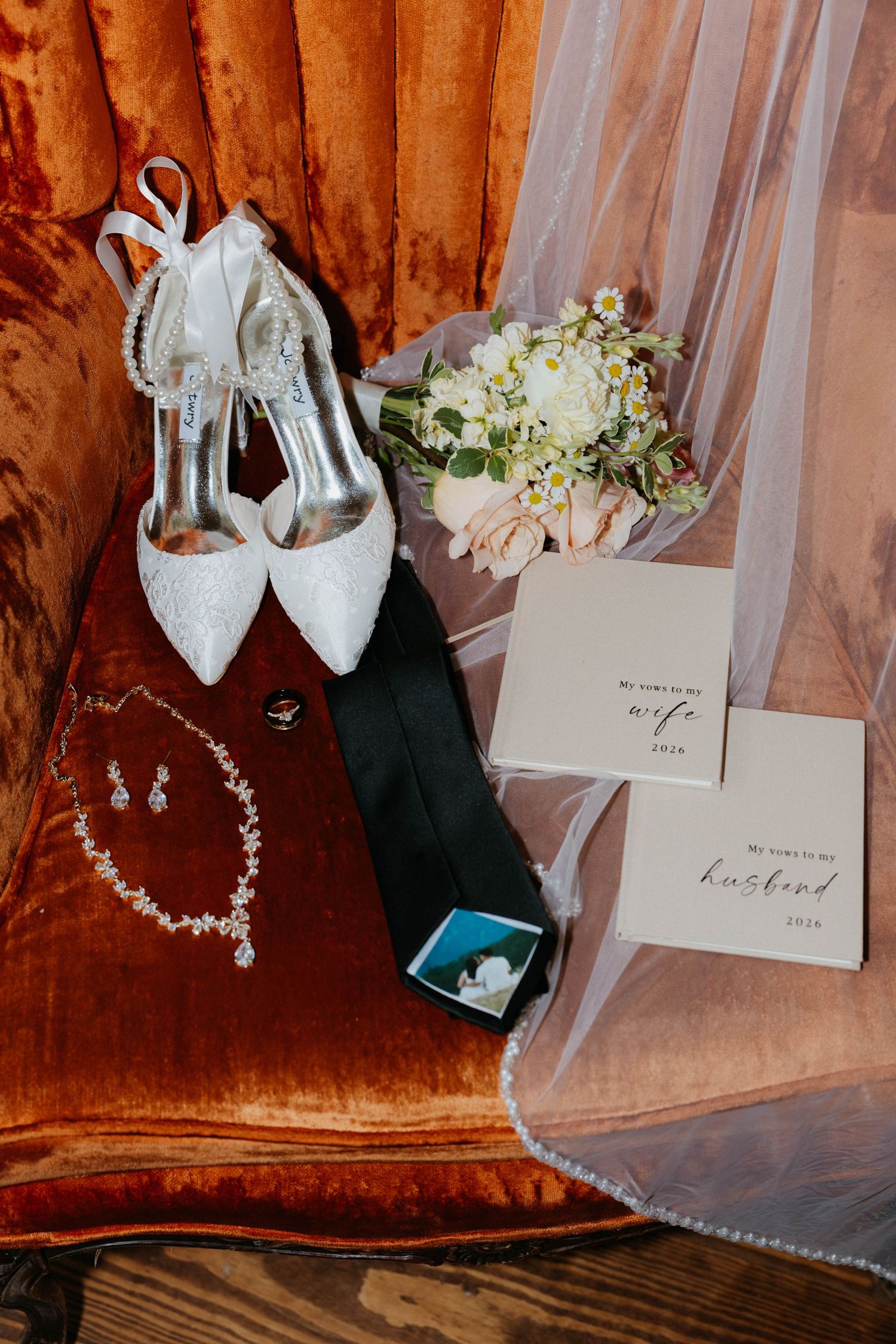 Elegant wedding flat lay featuring white lace heels, floral bouquet, veil, jewelry, black tie, and vow cards on velvet chair.