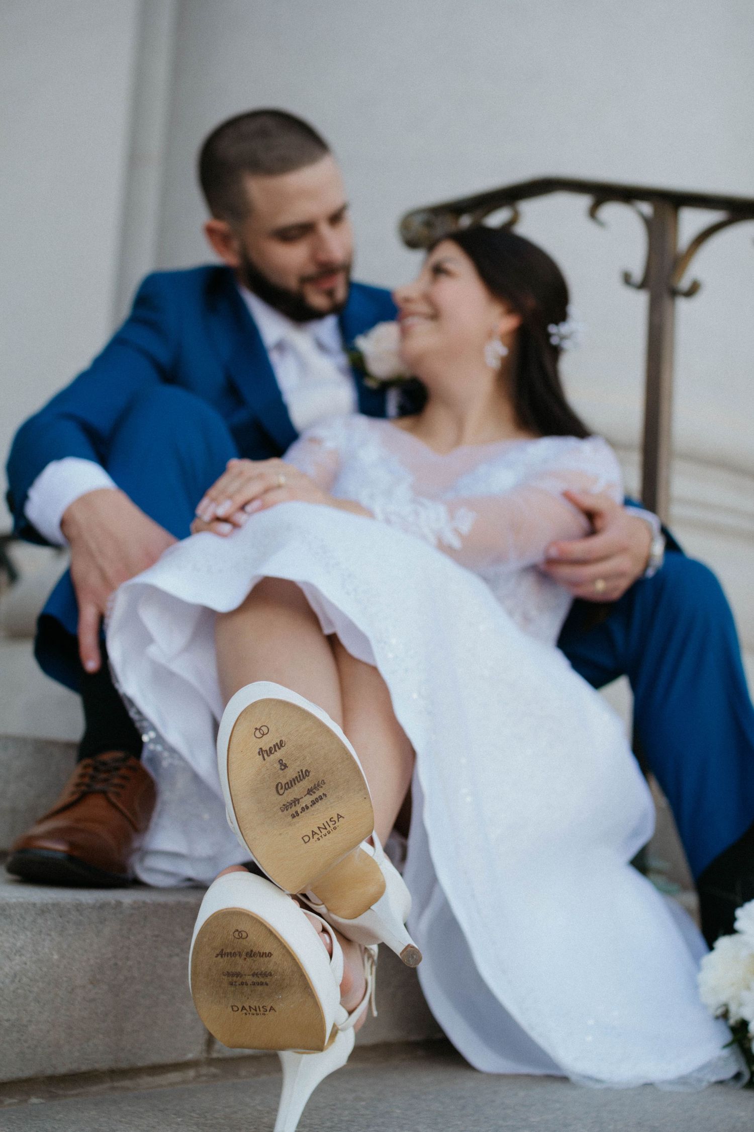 Bride and groom sitting on steps, bride's white heels with custom sole inscription visible in foreground.