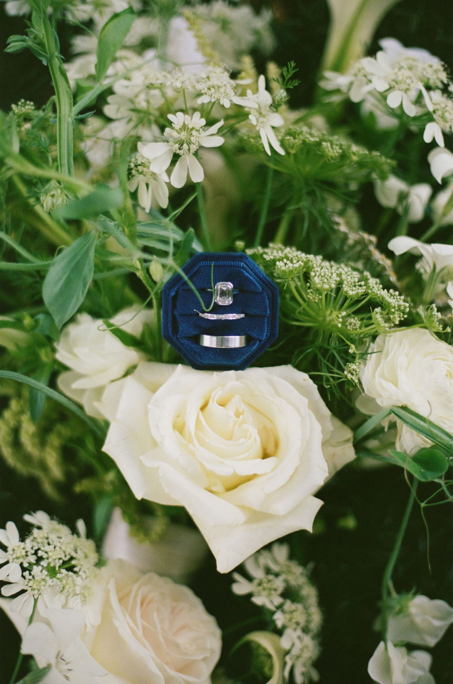 Wedding rings in a navy blue velvet box resting on white roses and wildflowers in a bridal bouquet.