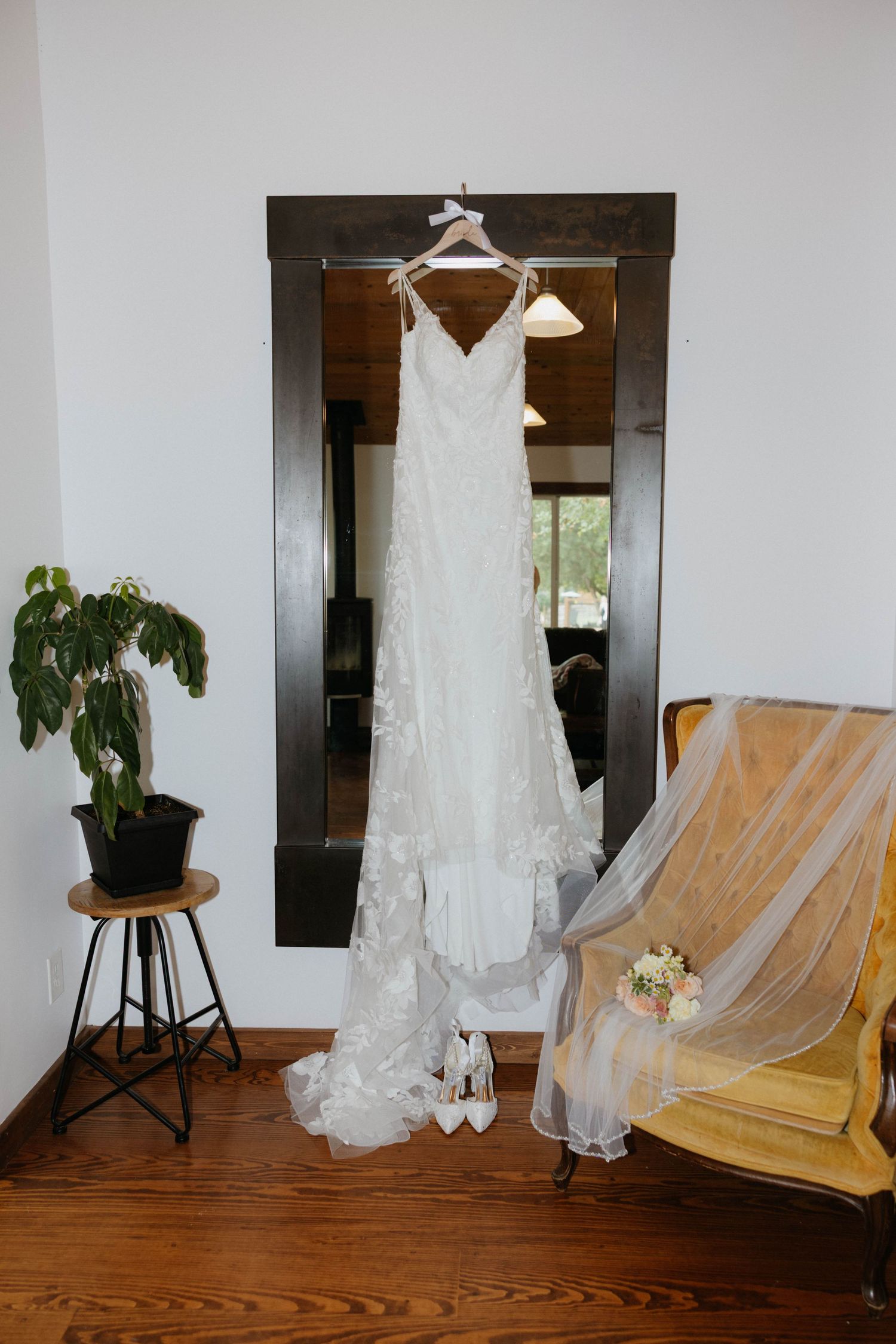 White lace wedding dress hanging on mirror frame with bridal heels and bouquet beside yellow chair.
