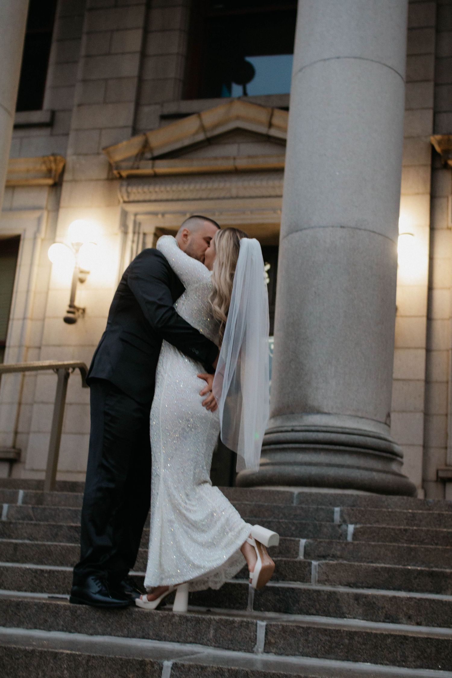A romantic wedding couple shares a dip kiss pose on stone steps with elegant columns in the background.