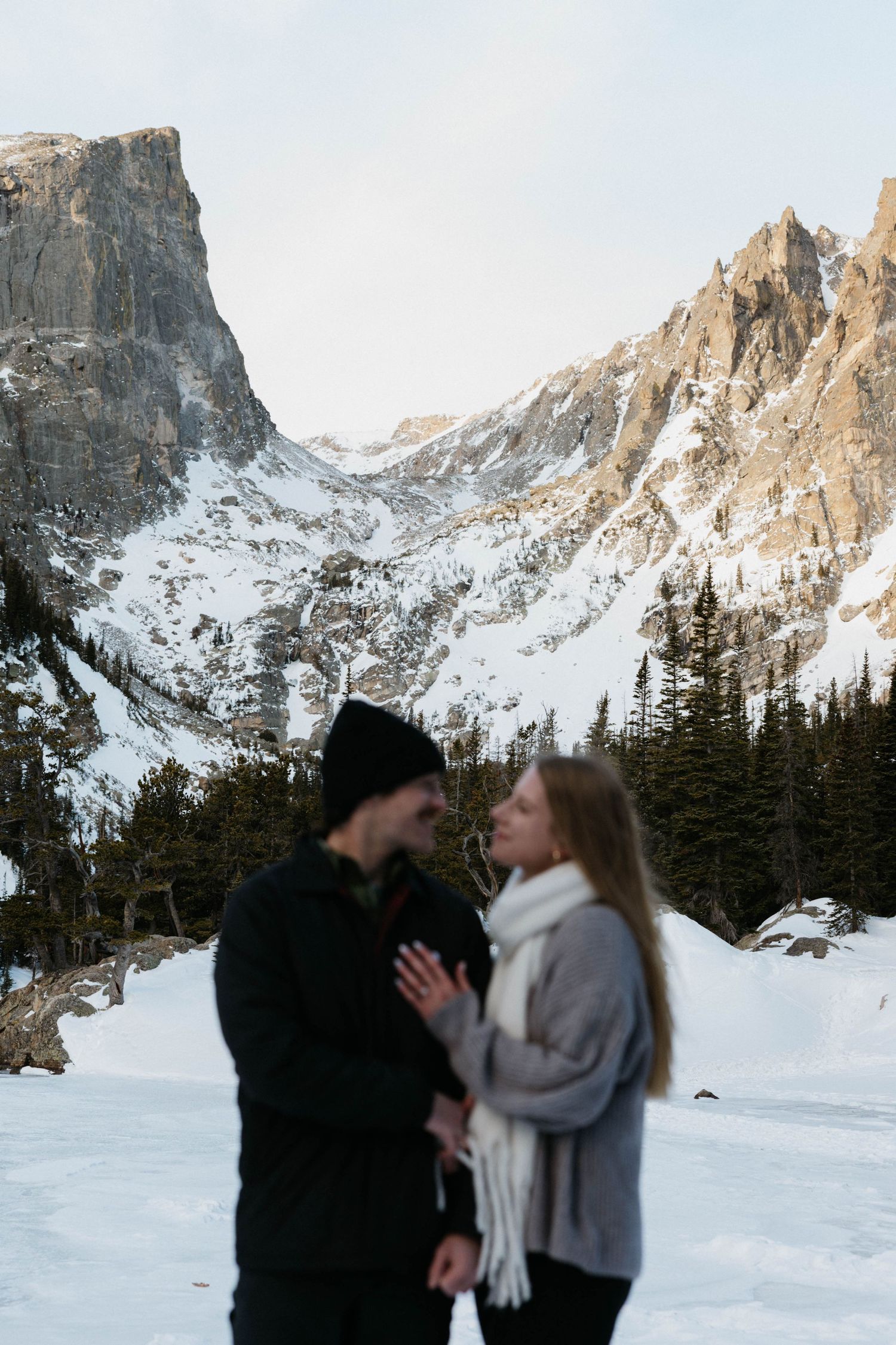 Couple sharing a romantic moment on a snowy mountain landscape with rocky peaks and evergreen trees in winter.