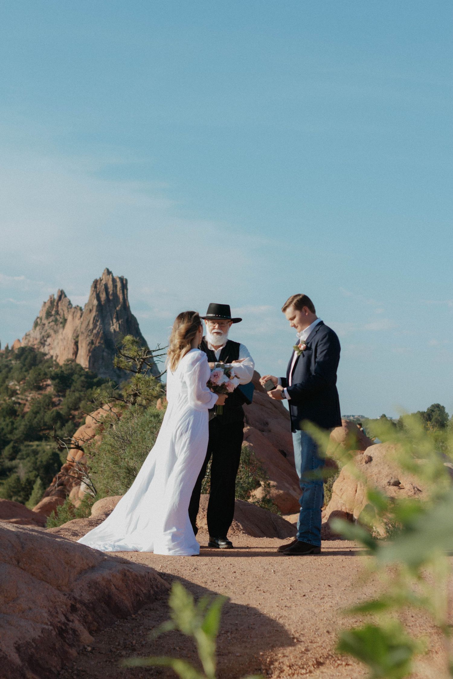 Outdoor wedding ceremony at Garden of the Gods, bride and groom exchanging vows with officiant, red rock formations backdrop.