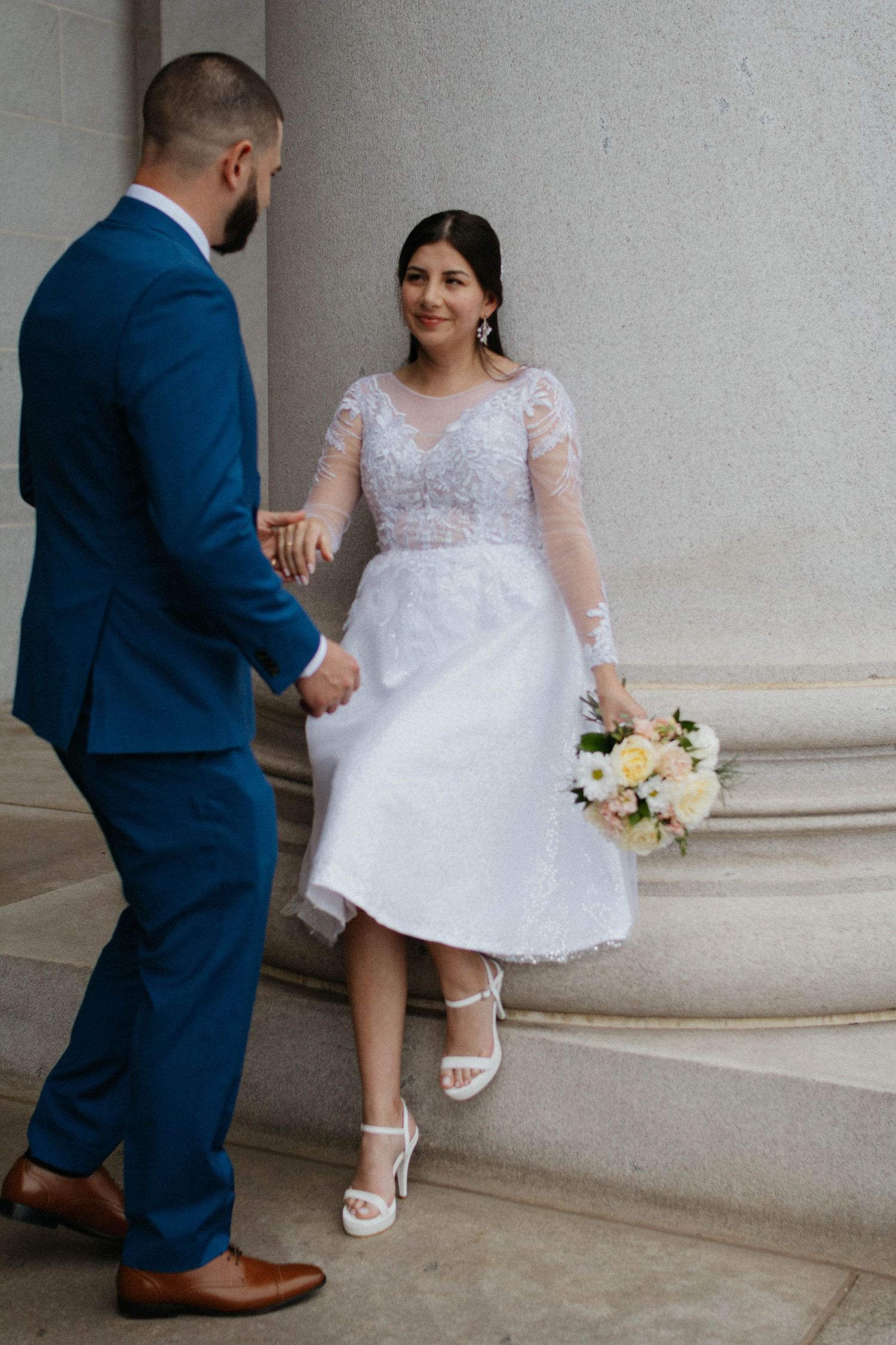 Bride in white lace midi dress holds bouquet while groom in blue suit holds her hand on stone steps.