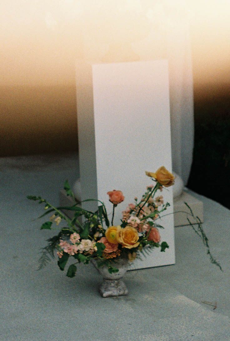 Elegant floral arrangement with yellow and peach roses in a stone urn beside a blank white card at golden hour.