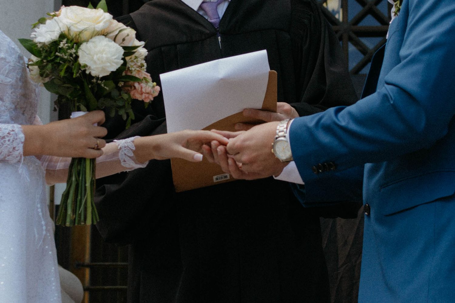 Groom in blue suit places wedding ring on bride's finger during outdoor ceremony, bride holding white floral bouquet.