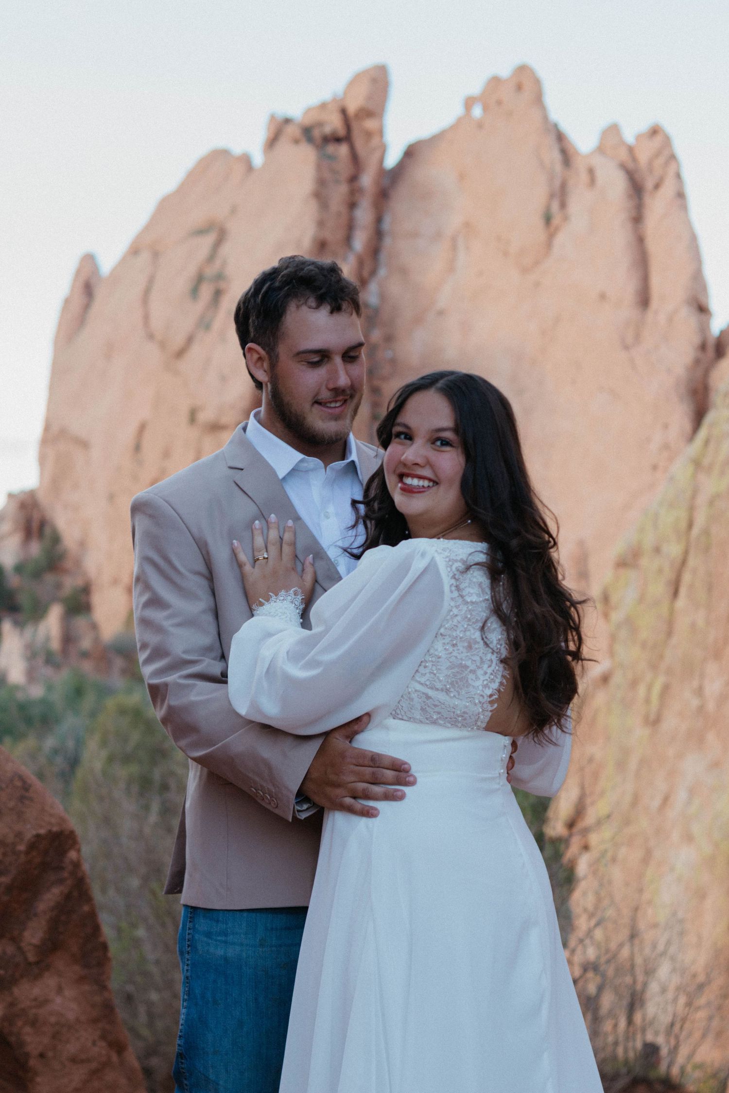 Couple embracing in white dress and tan blazer with red rock formations at Garden of the Gods Colorado backdrop.