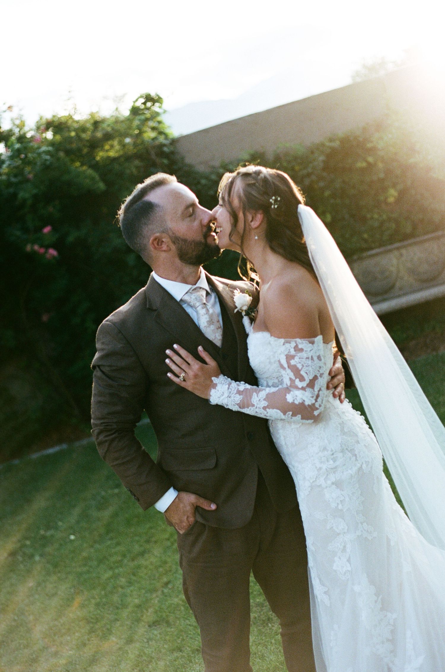 A couple shares a romantic almost-kiss in golden sunlight, bride in lace gown with veil, groom in dark suit.