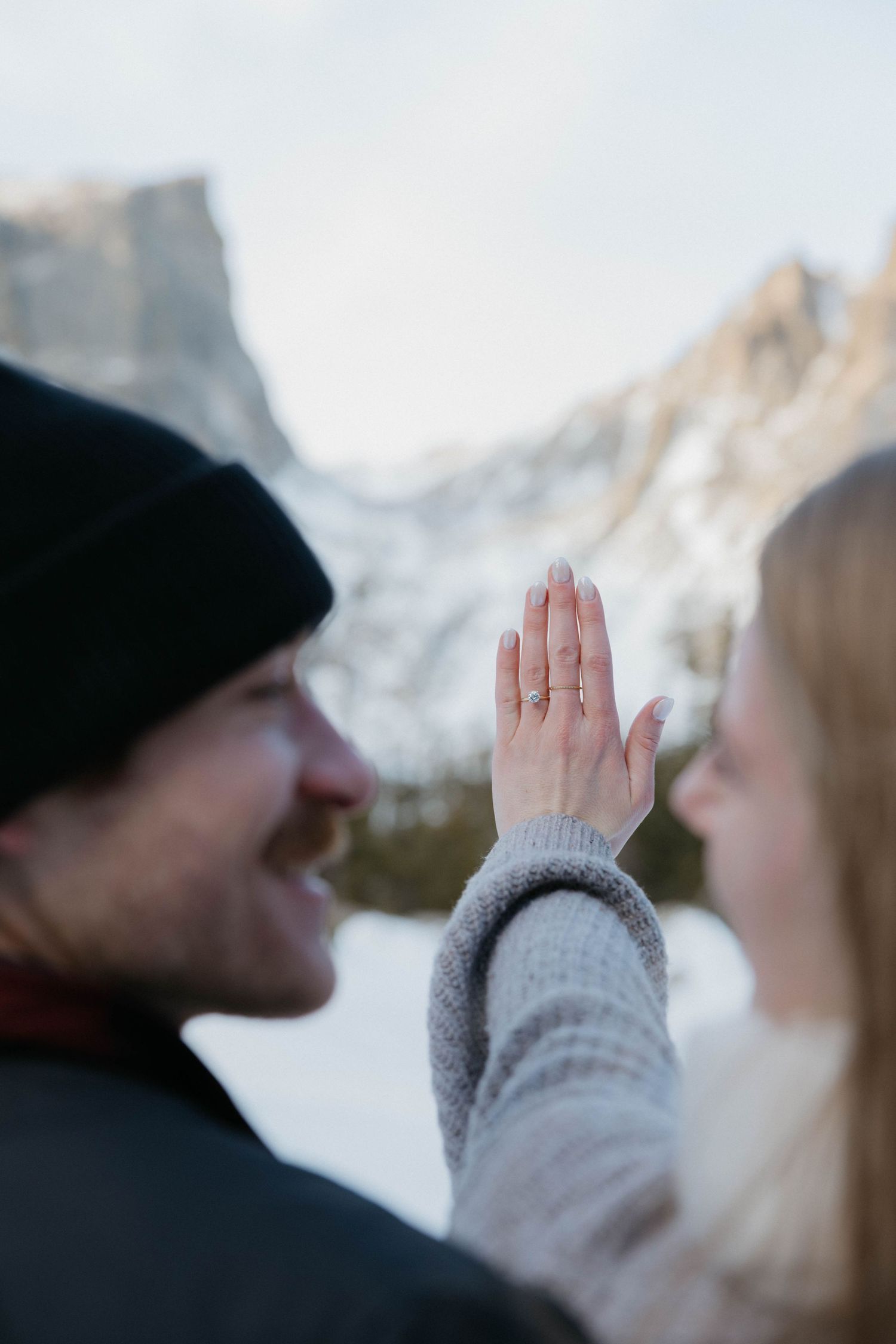 Couple sharing a tender moment in snowy mountains, woman showing engagement ring on outstretched hand.