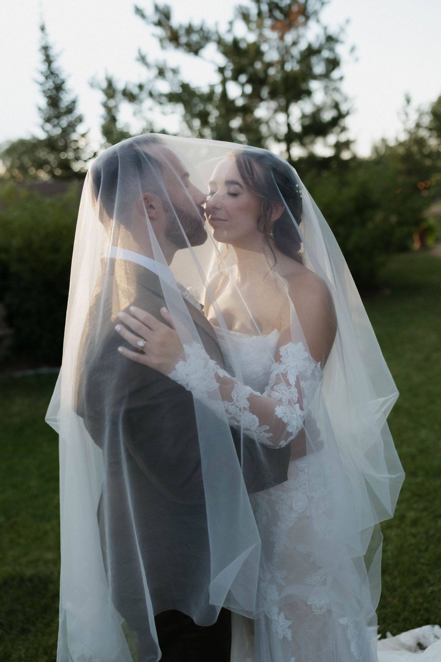 Bride and groom sharing a romantic kiss under a flowing white veil outdoors on their wedding day.