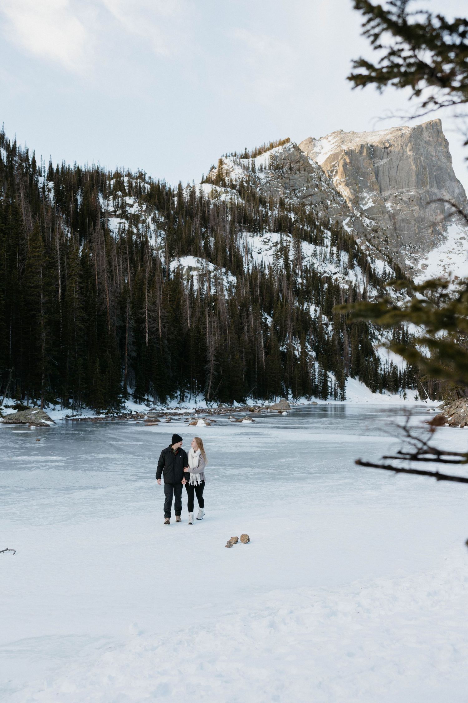 Couple holding hands on a frozen lake surrounded by snow-covered pine trees and rocky mountains in winter.