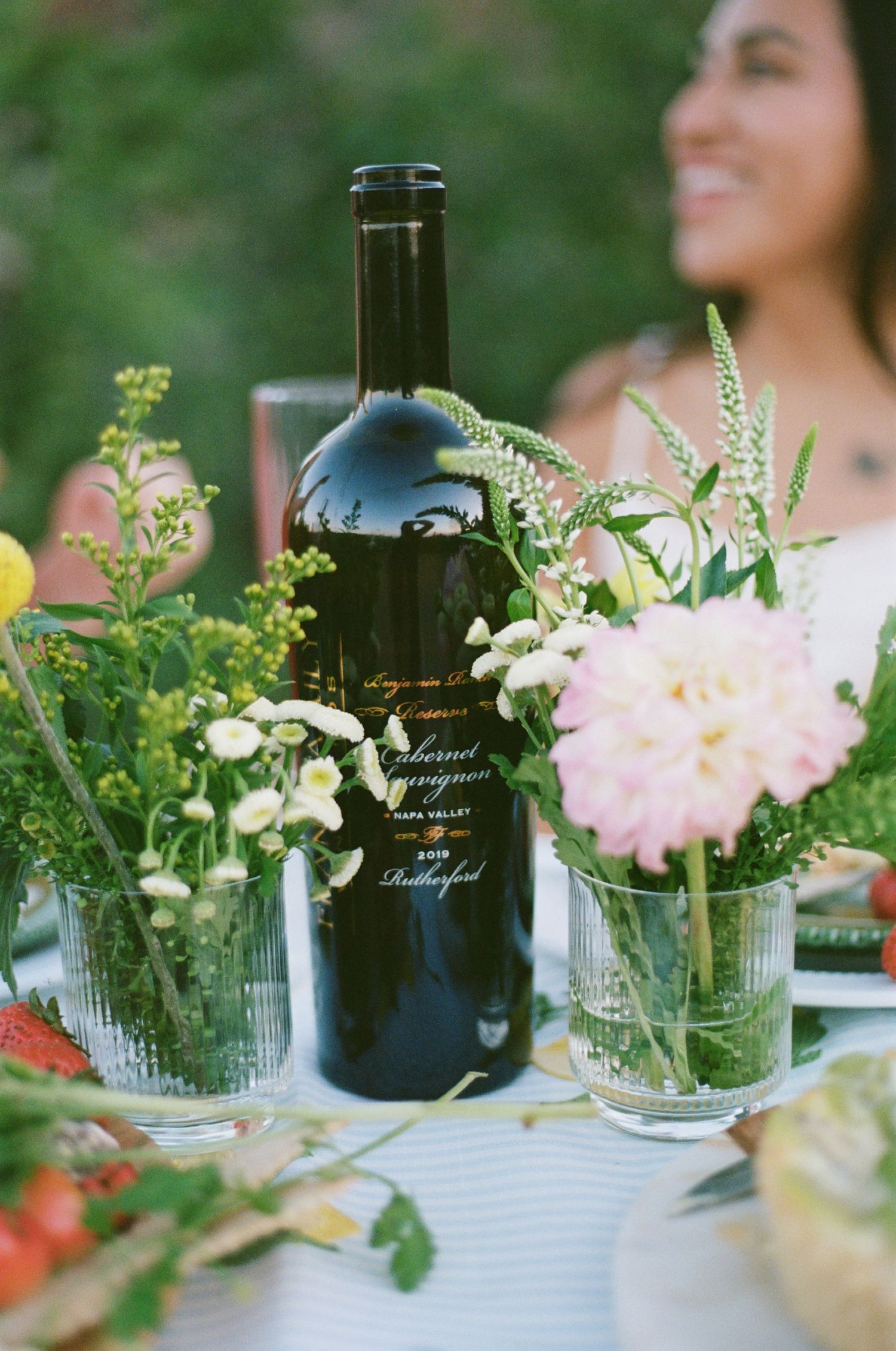 A bottle of Cabernet Sauvignon wine surrounded by fresh flowers in glass vases at an outdoor garden party table.