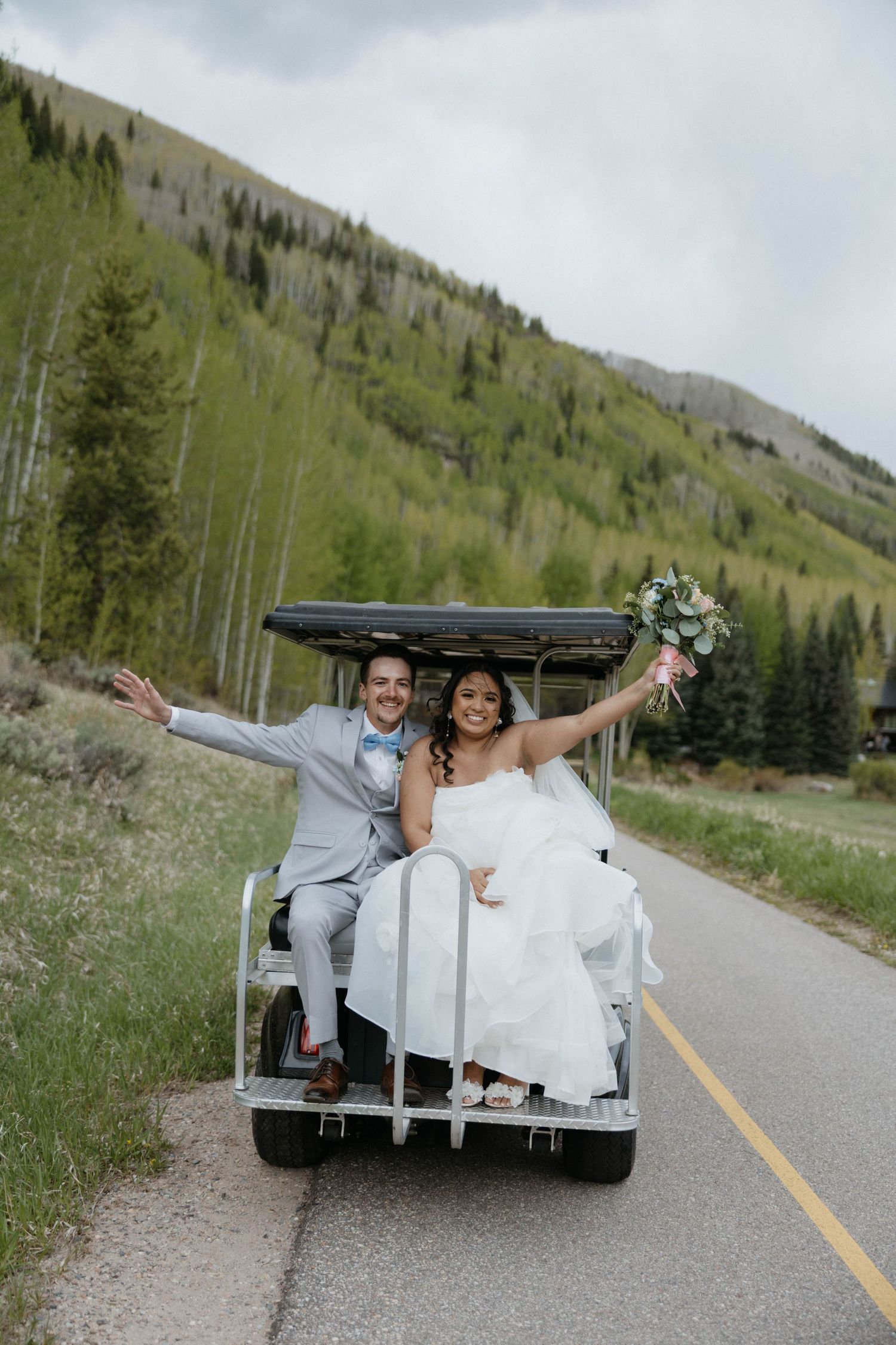 Bride and groom celebrate on a golf cart on a mountain road surrounded by lush green trees in Colorado.