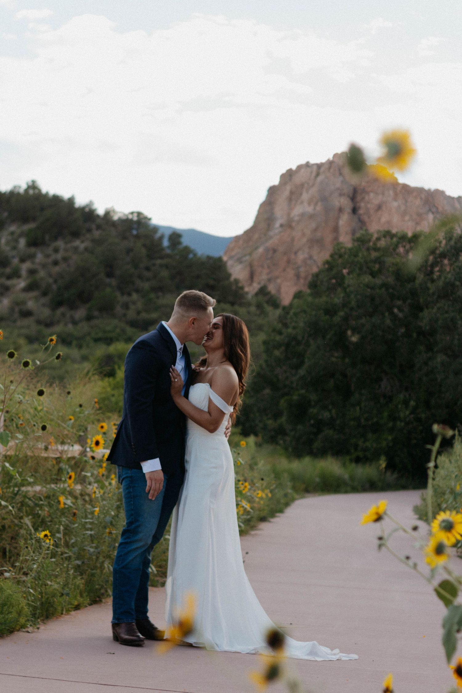 Bride and groom share a romantic kiss on a garden path with sunflowers and red rock formations in Colorado.
