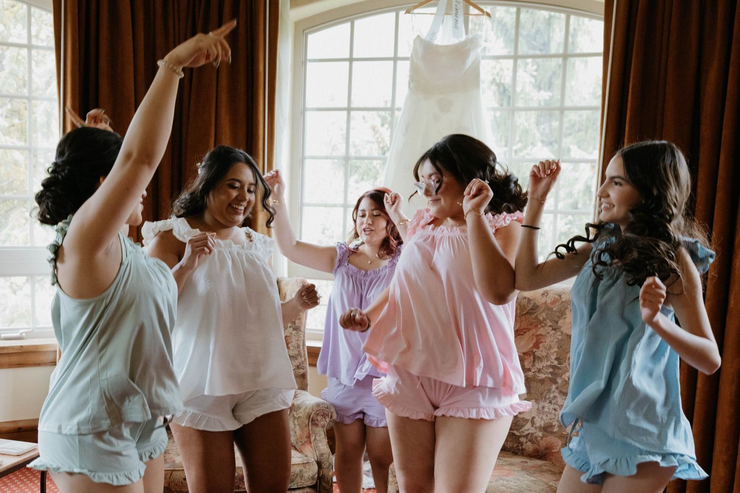 Bridesmaids in pastel pajamas dancing joyfully in a sunlit room with a wedding dress hanging in the background.
