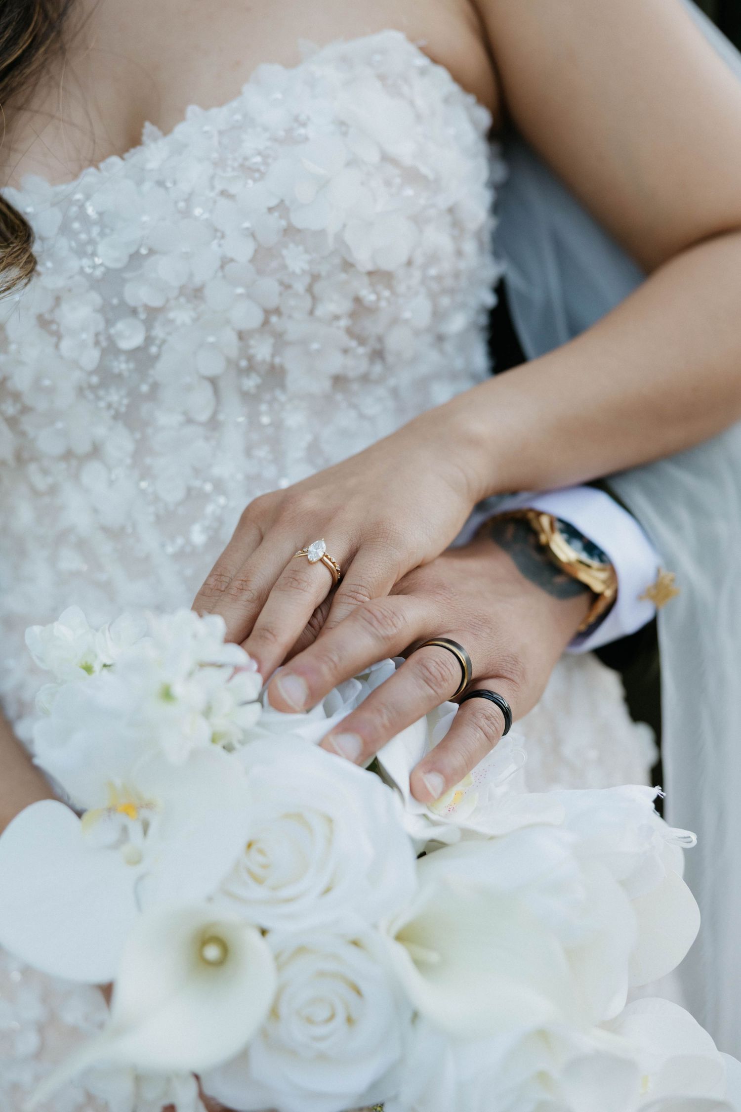 Bride and groom hands with wedding rings resting on white floral bouquet with roses and calla lilies.