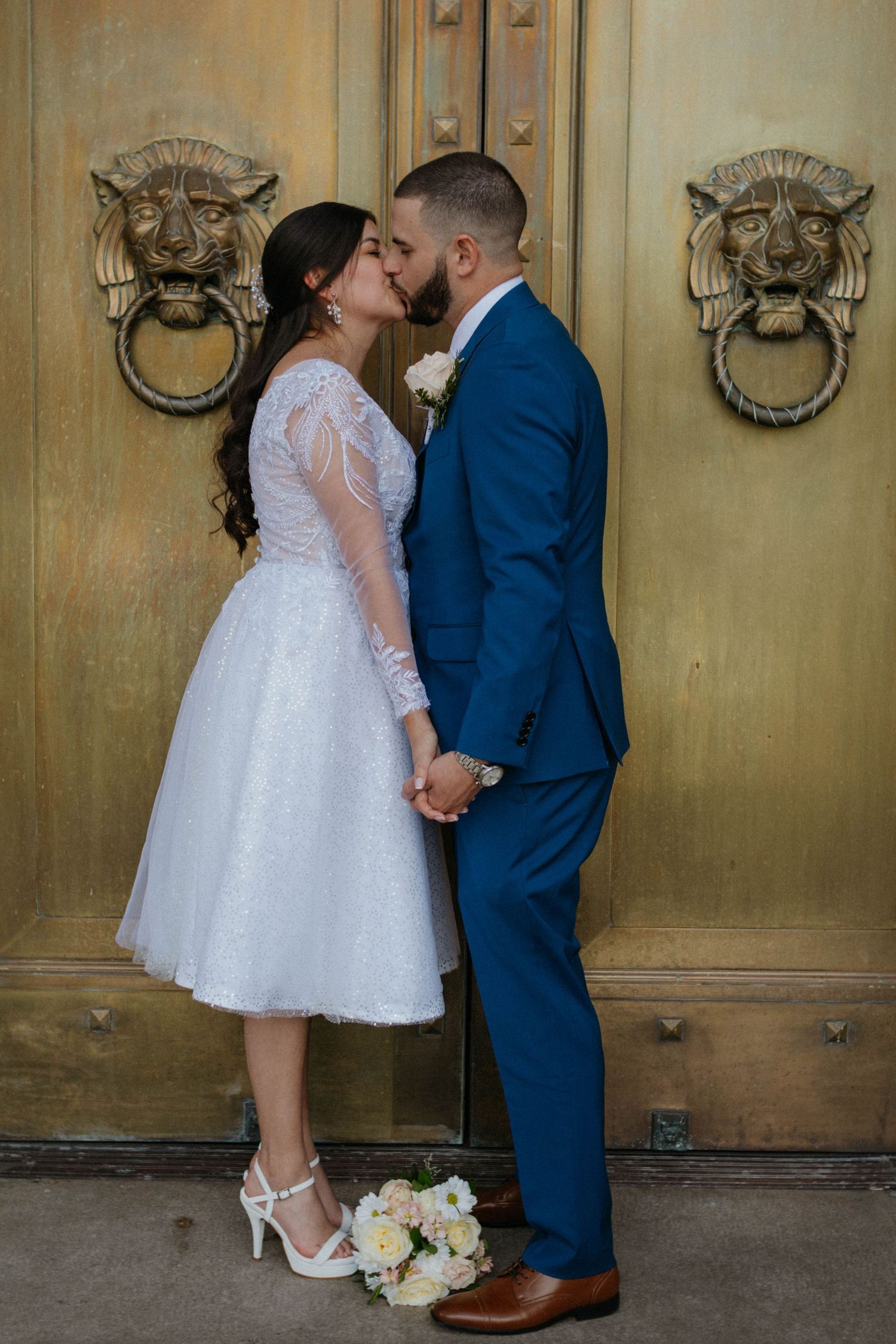 Newlywed couple kissing in front of golden doors with lion knockers, bride in white lace dress, groom in blue suit.