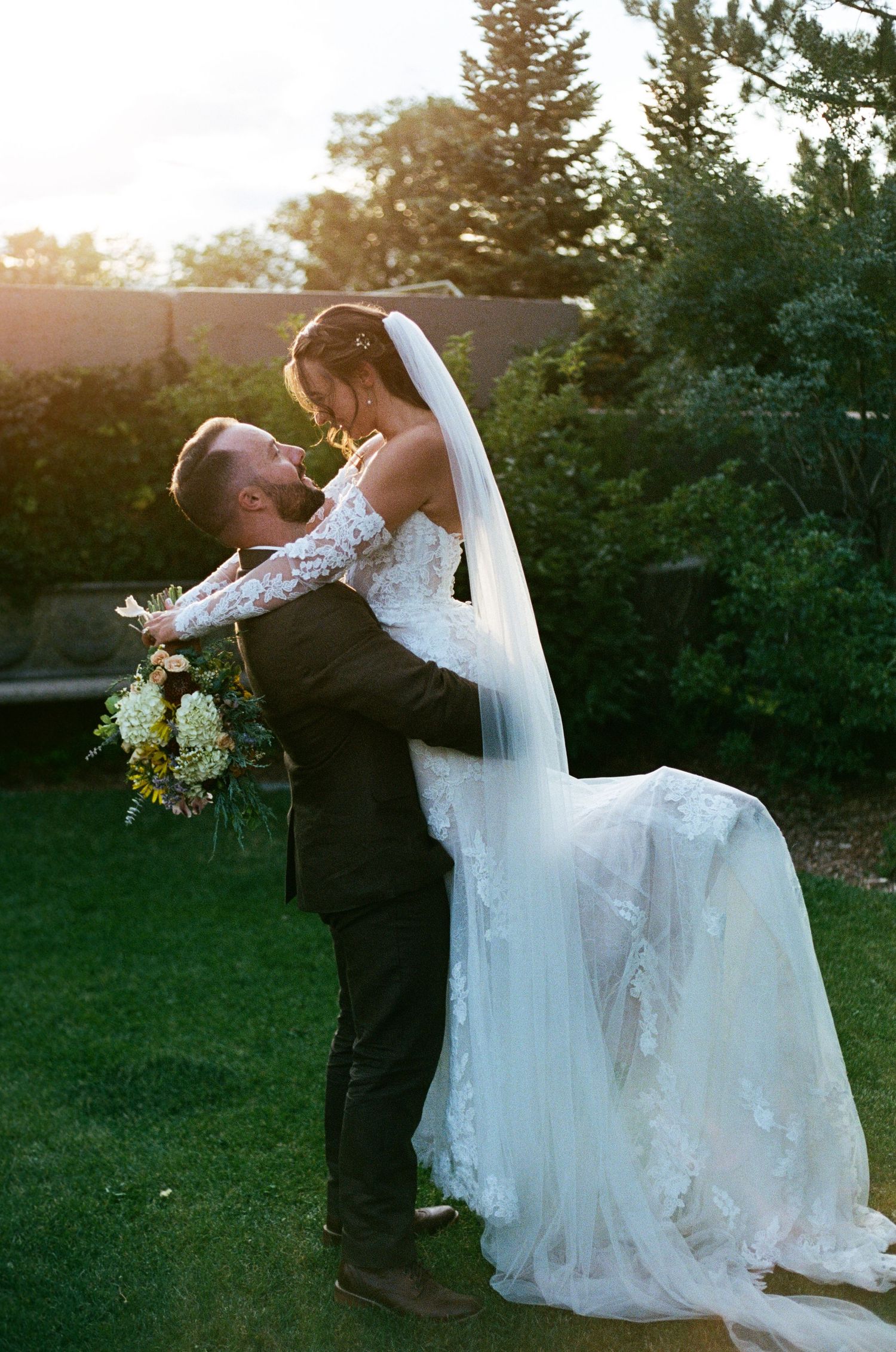 Groom lifts bride in lace wedding dress and veil outdoors at golden hour, surrounded by lush greenery.