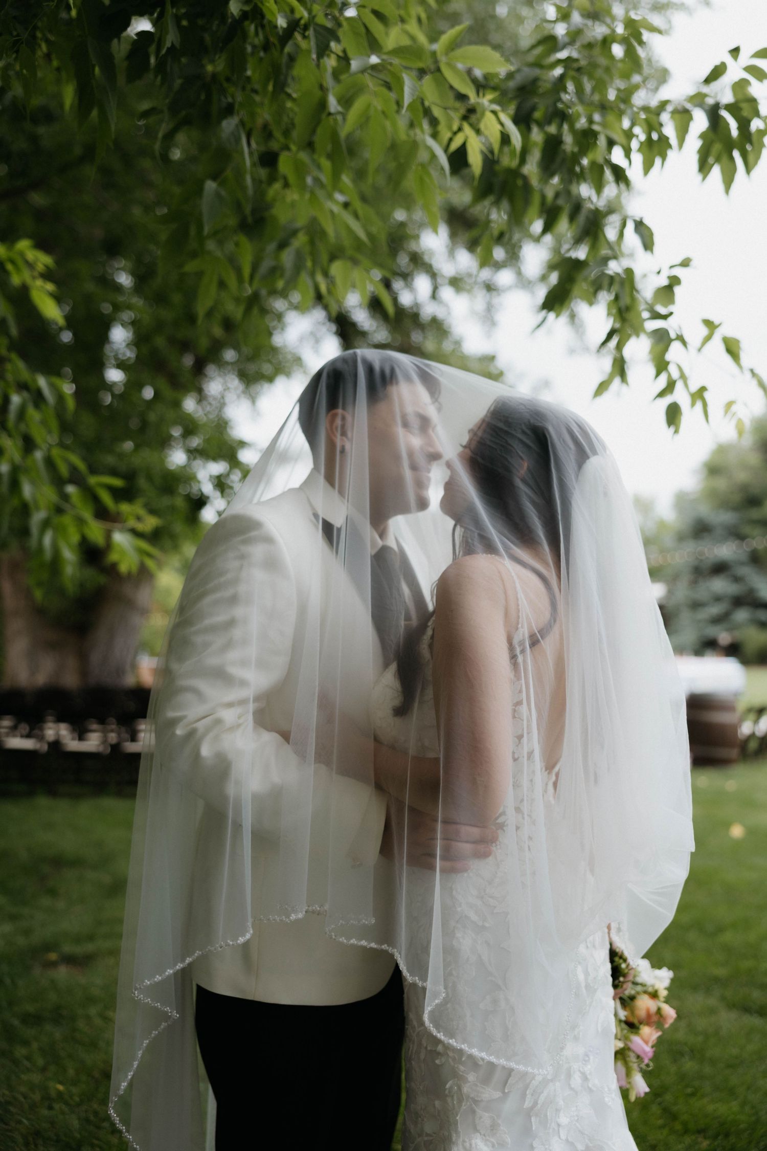 Bride and groom kissing under a white veil outdoors, surrounded by lush green trees on their wedding day.