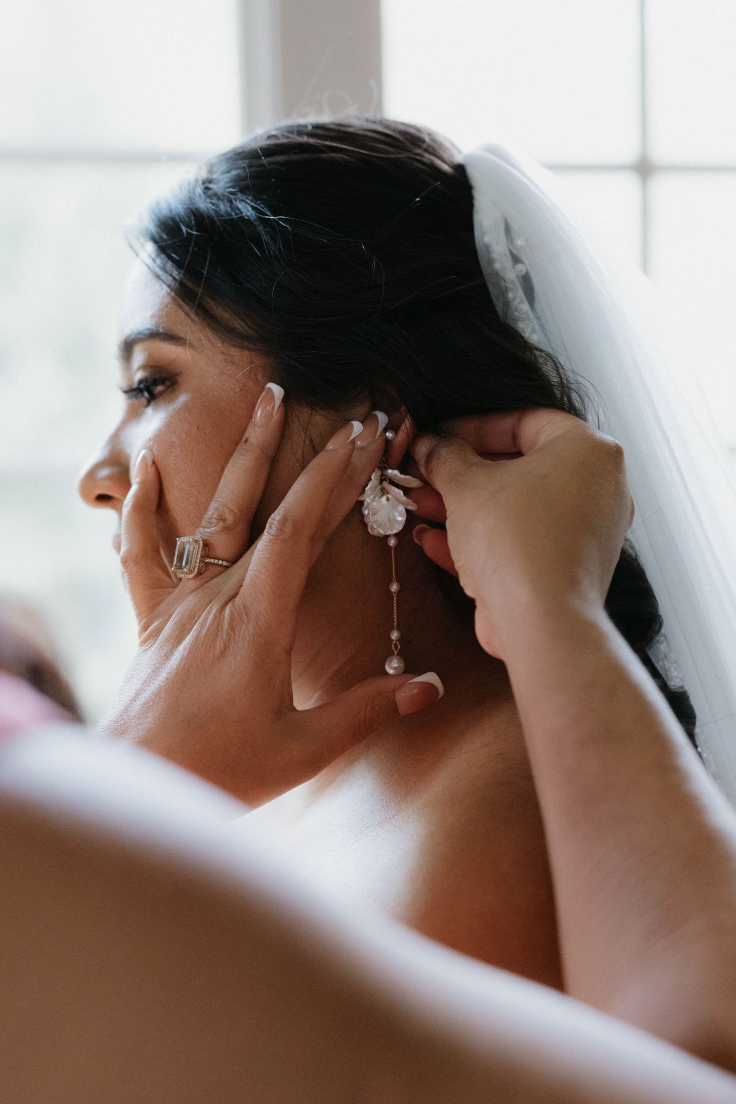 A bride wearing a veil has elegant drop earrings put on by a helper during wedding preparations.