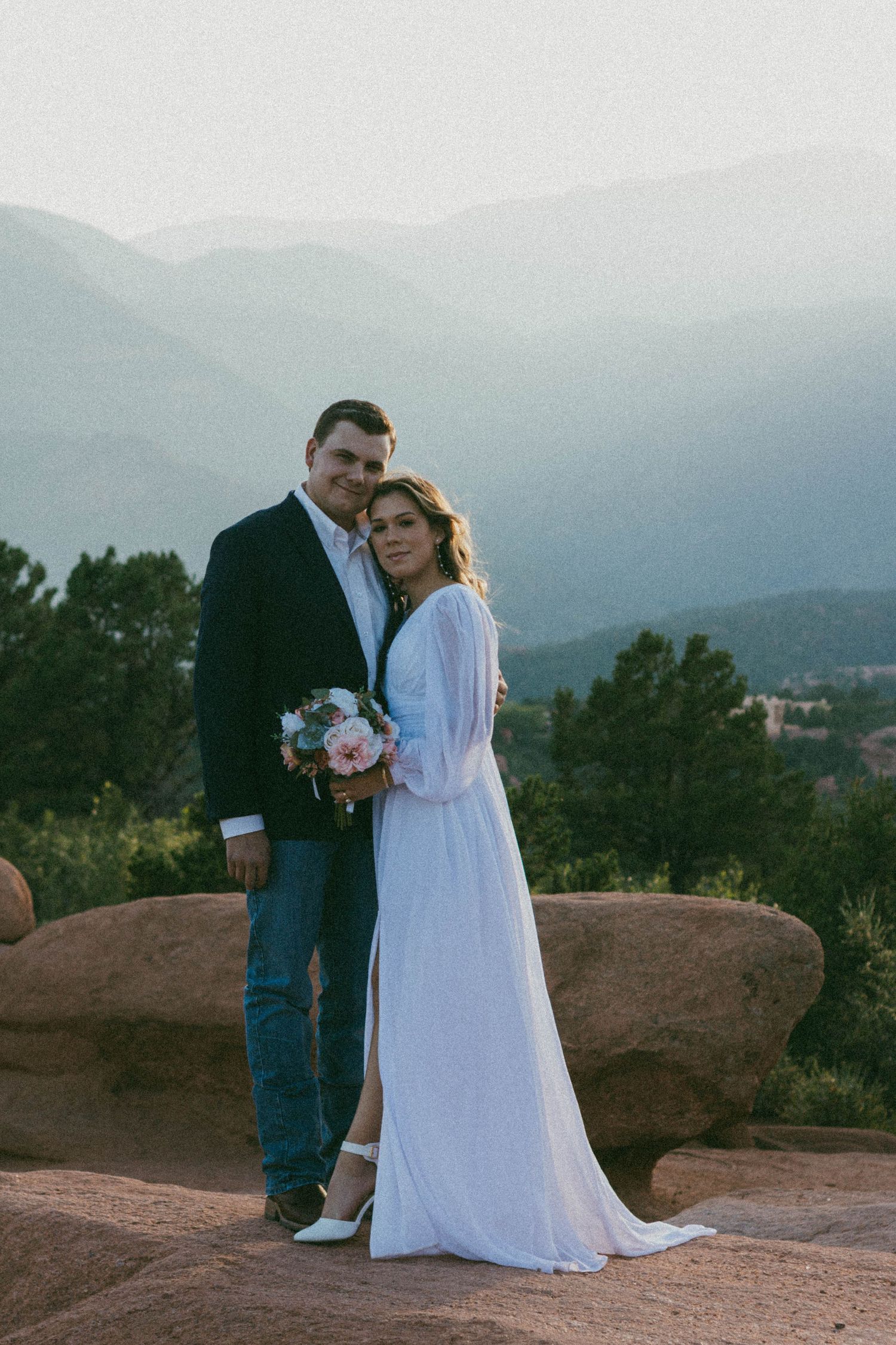 Couple in wedding attire poses on rocky overlook with mountain views, bride in white gown holding bouquet.