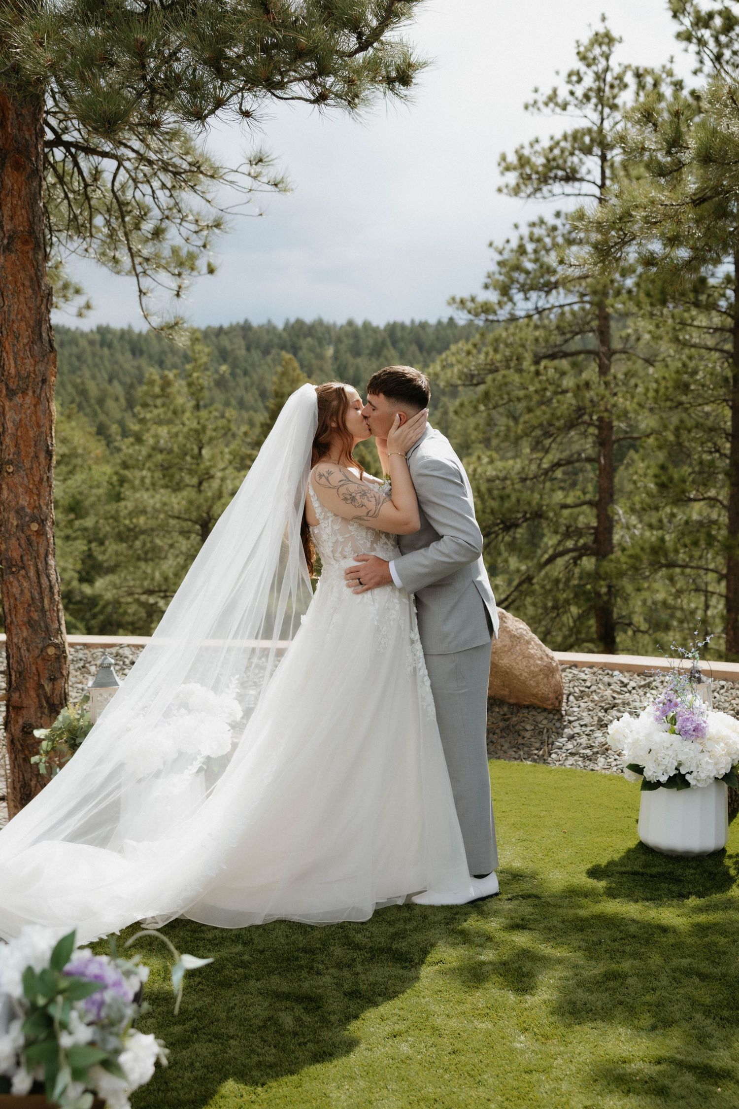 Bride and groom share their first kiss at an outdoor wedding ceremony surrounded by pine trees and mountain views.