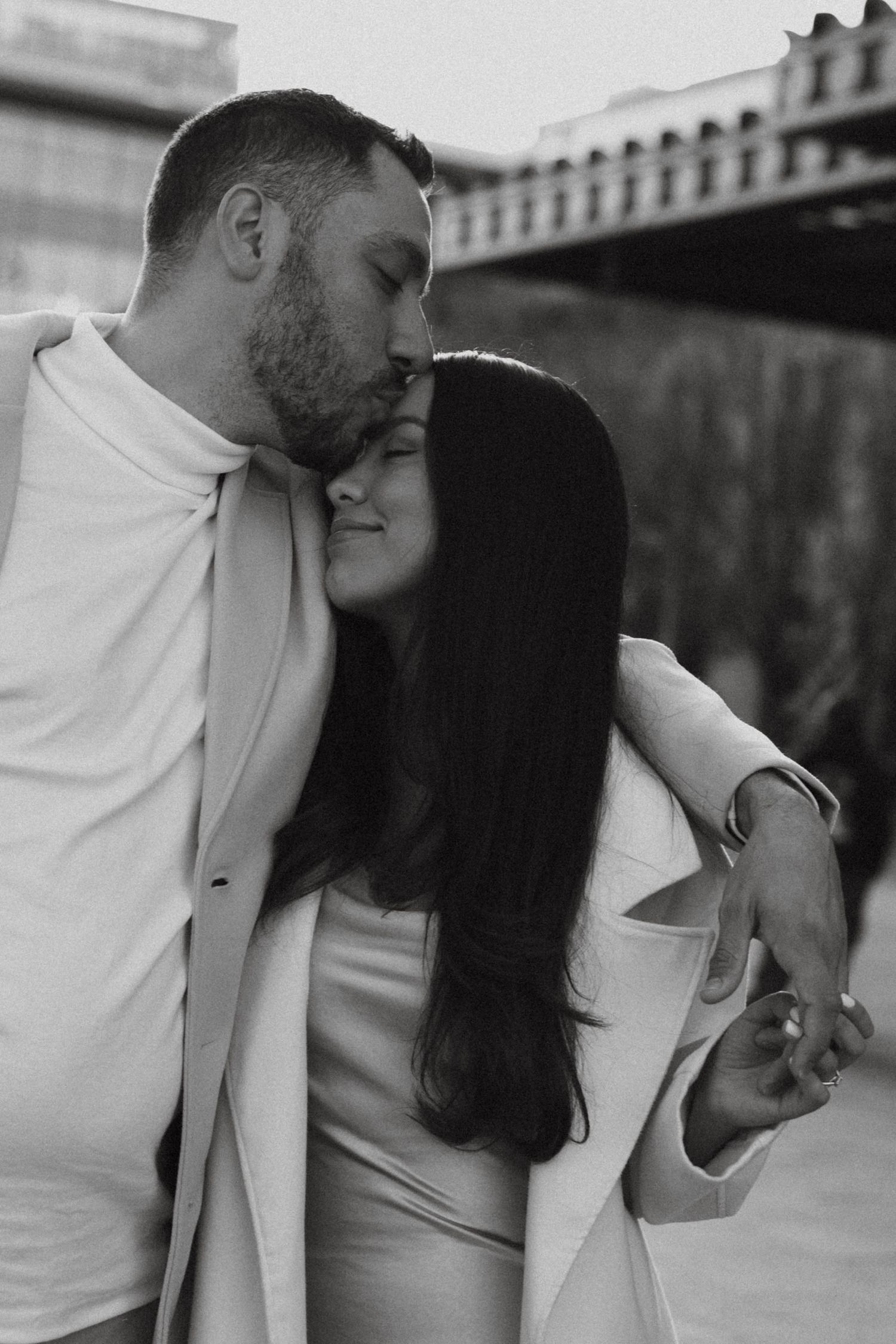 Black and white photo of a couple embracing, man kissing woman on the head outdoors near a bridge.