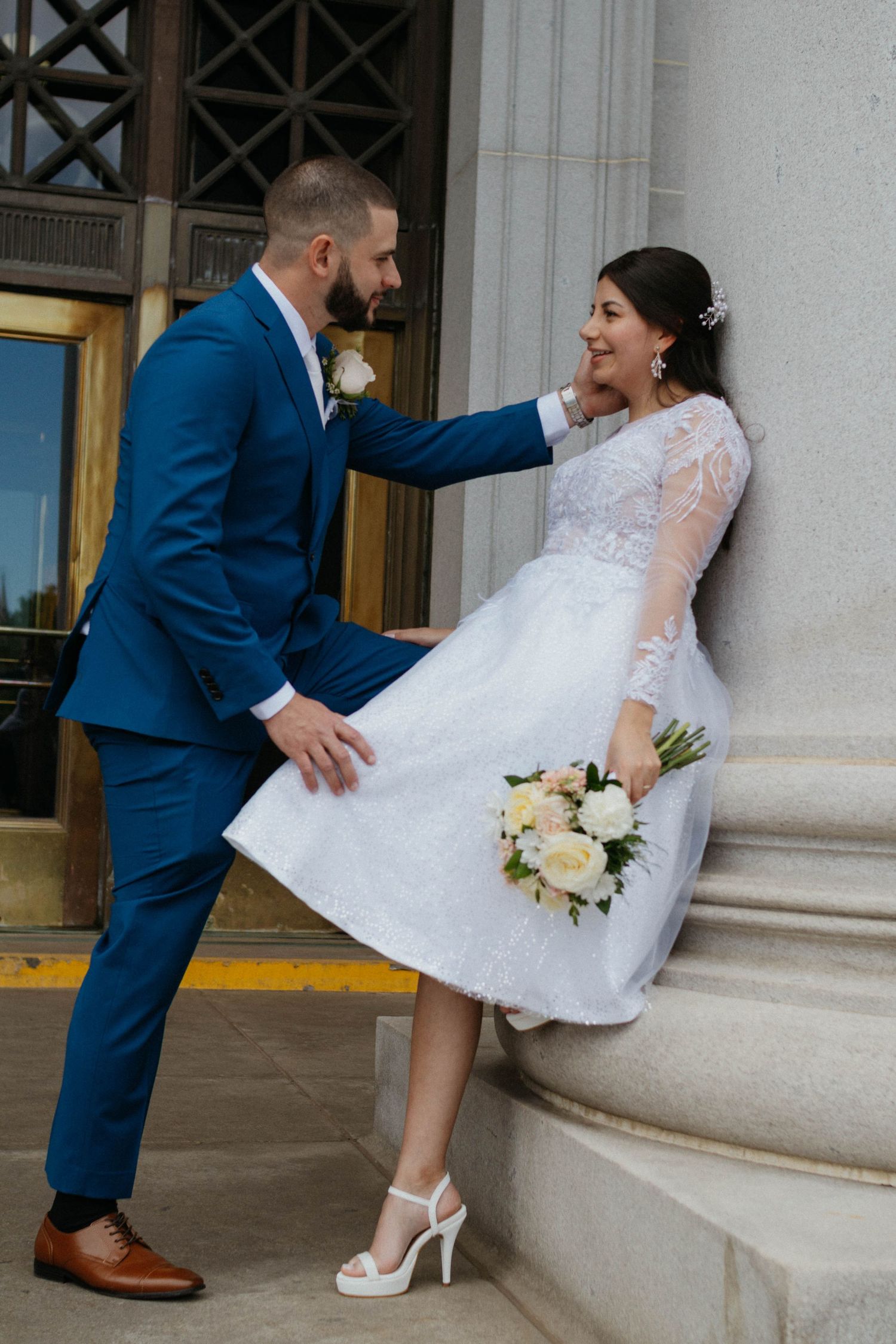 A groom in a blue suit gazes at his bride in a white lace dress holding a bouquet by a stone column.