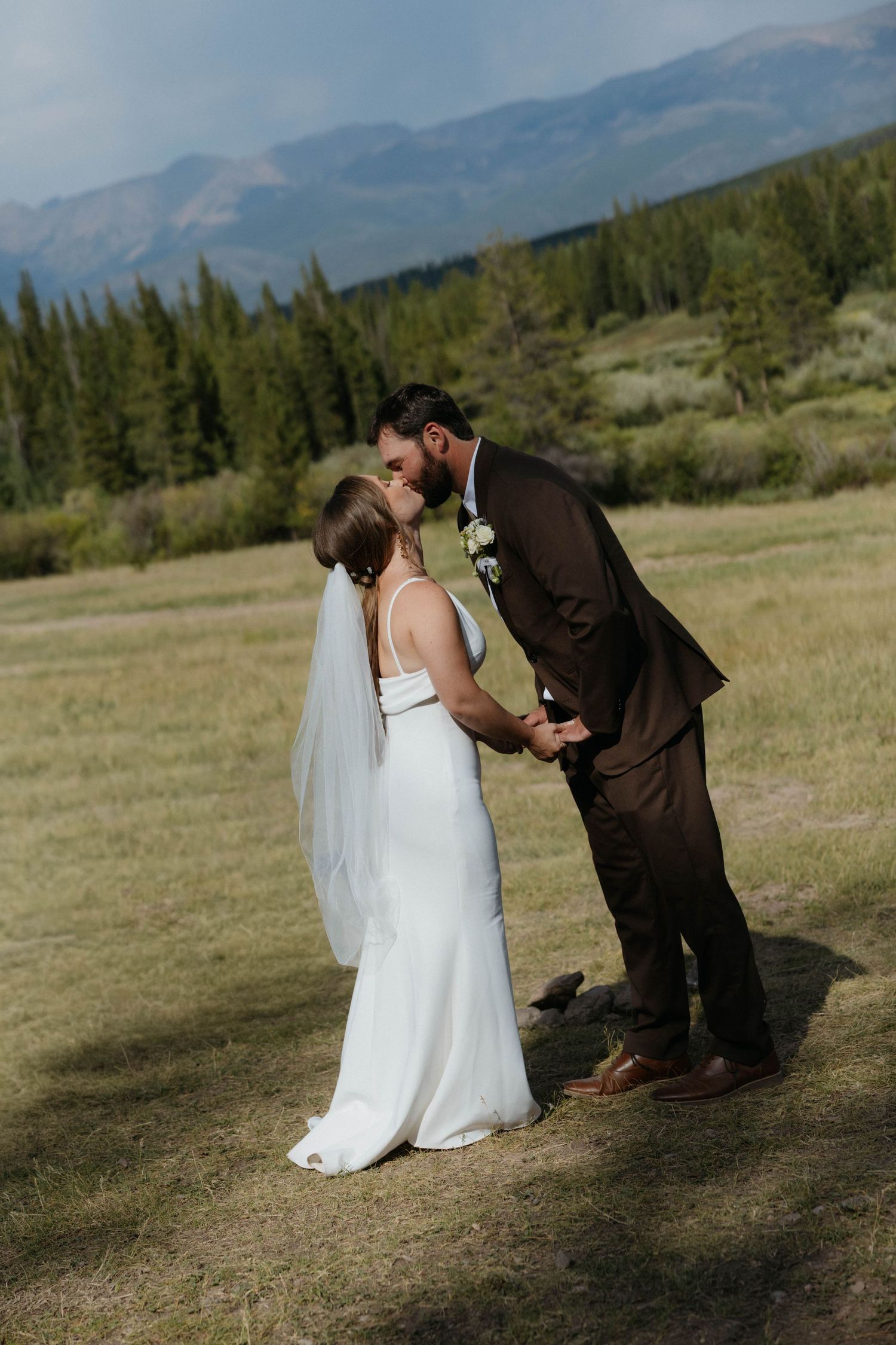 Bride and groom share a kiss in a mountain meadow, surrounded by pine trees, bride in white gown with veil.