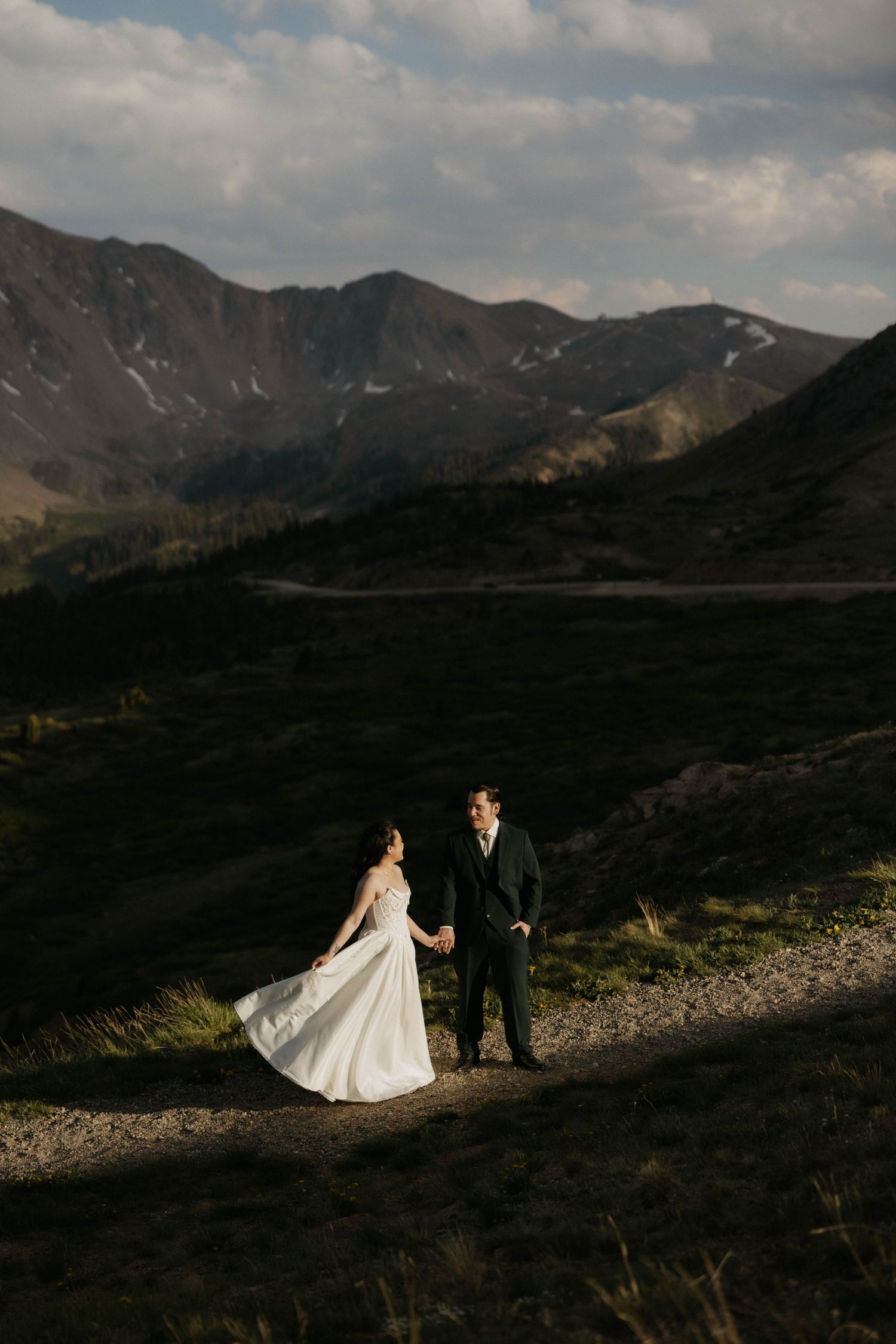 Bride and groom hold hands on a mountain trail at sunset with scenic Colorado peaks in the background.