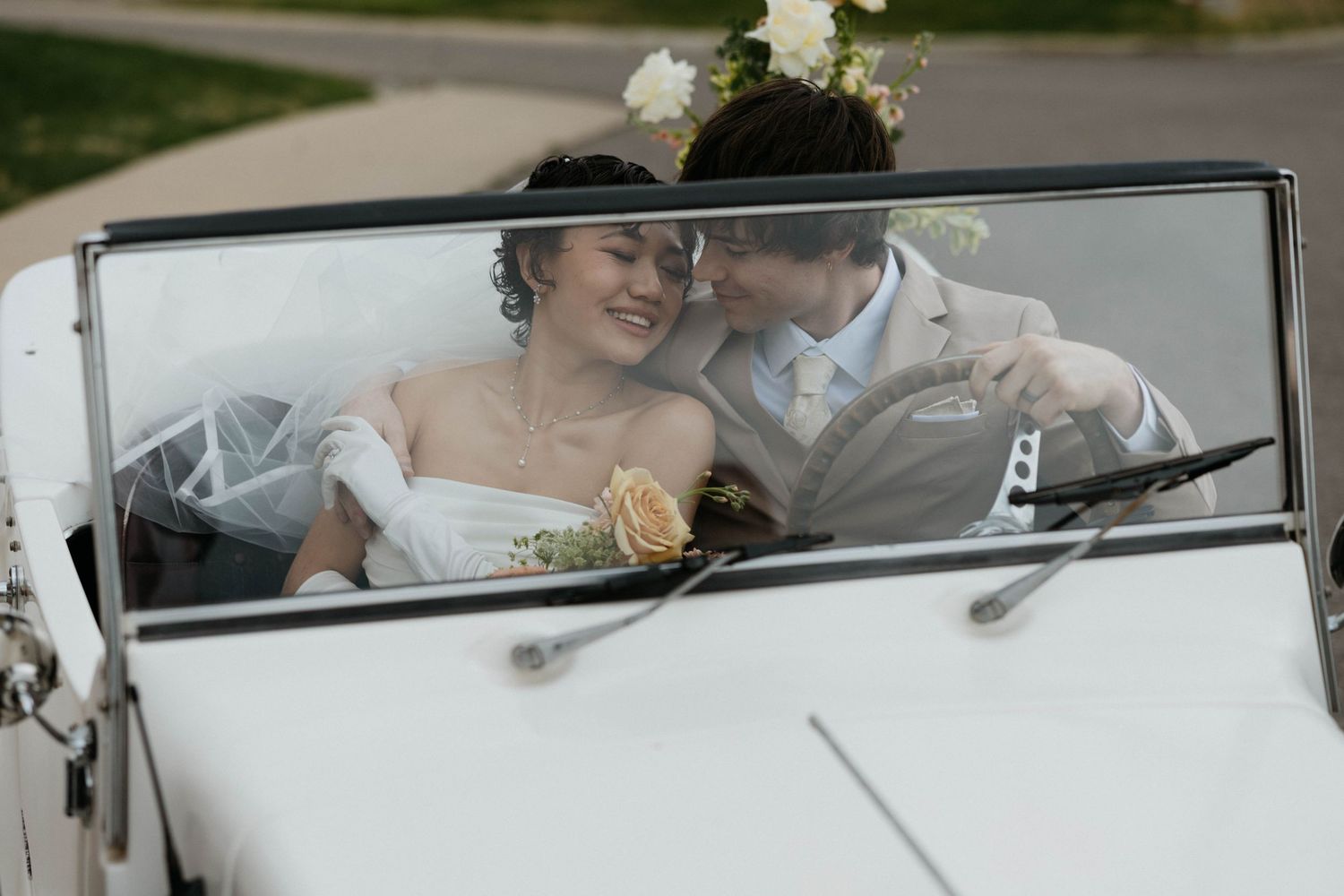Bride and groom smiling together in a white vintage convertible car on their wedding day.