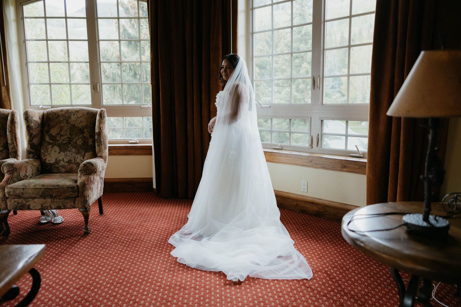 Bride in a white wedding gown with long veil stands by large windows in an elegant hotel room.