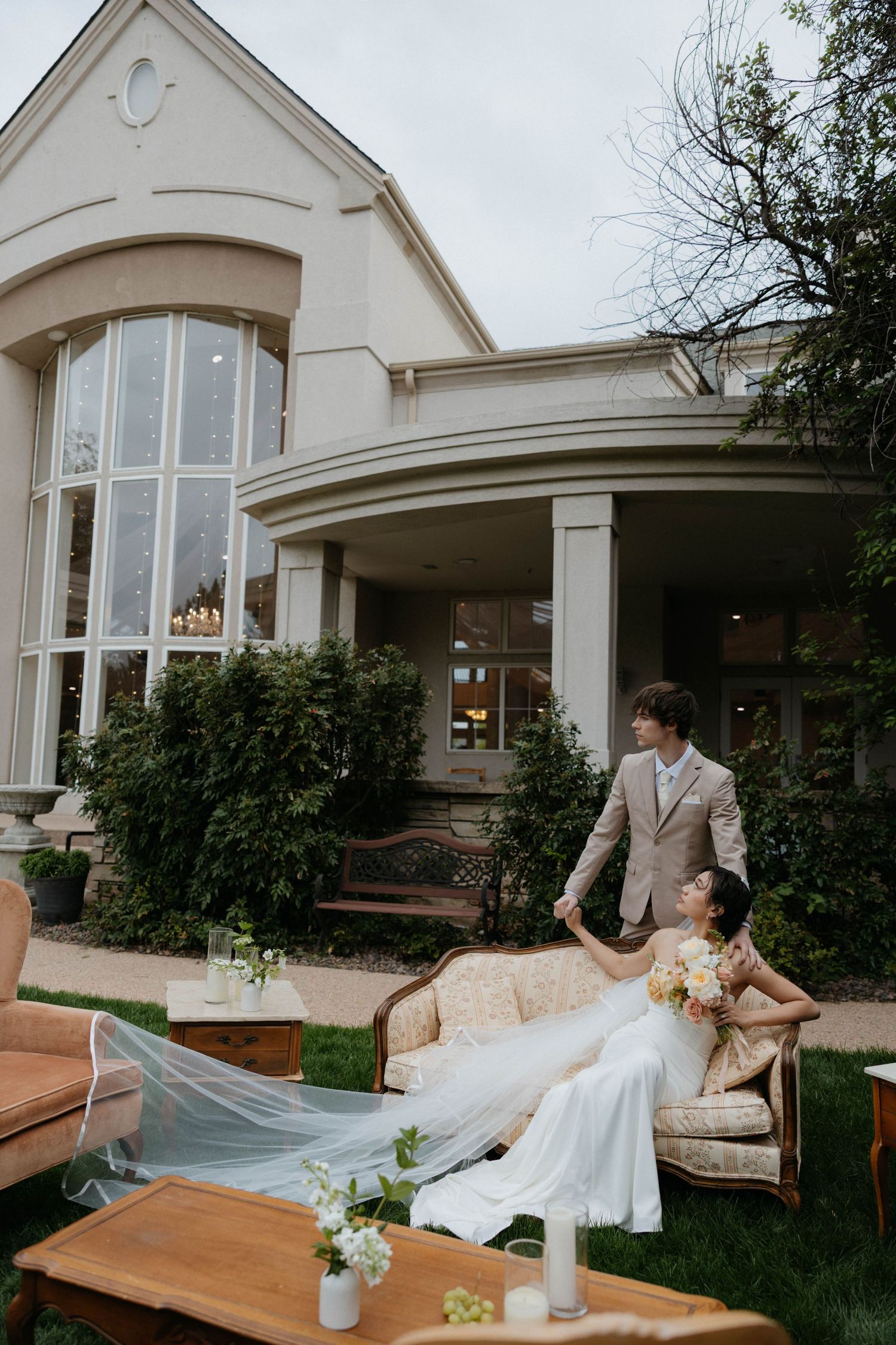 A bride in a flowing white gown sits on a vintage sofa outdoors while her groom in a tan suit stands beside her.
