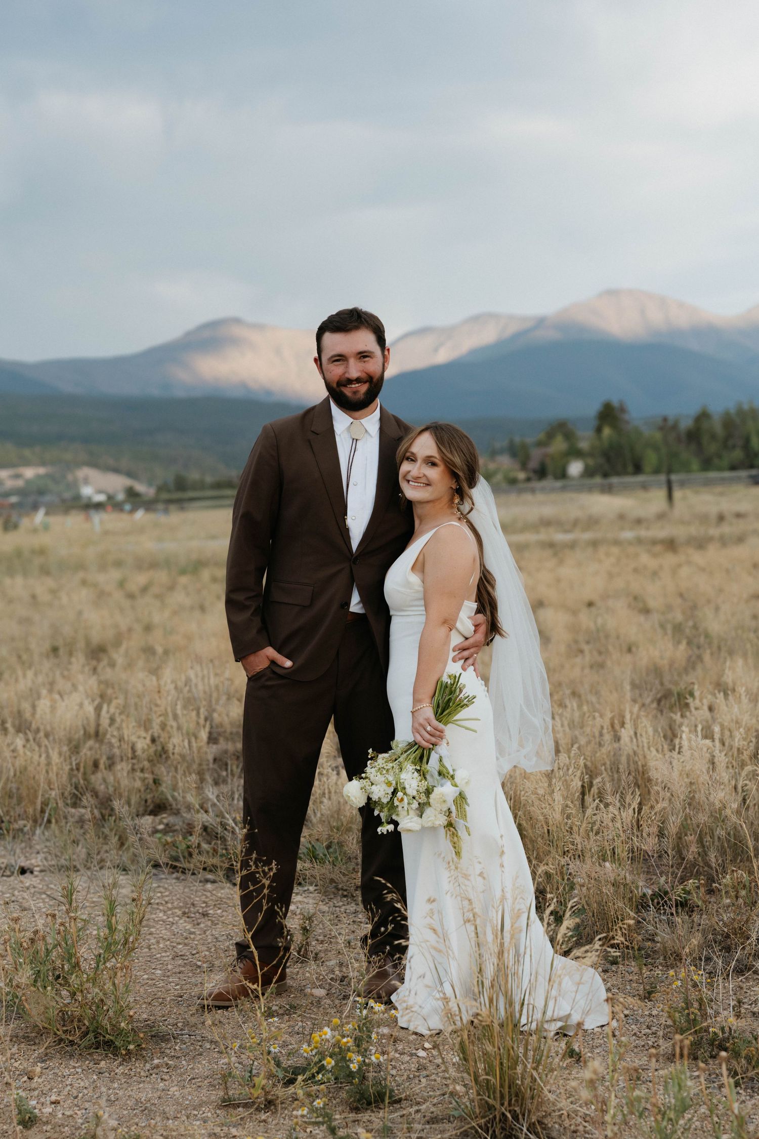 Bride and groom pose in a mountain meadow, she holds white bouquet and wears veil, he wears brown suit.