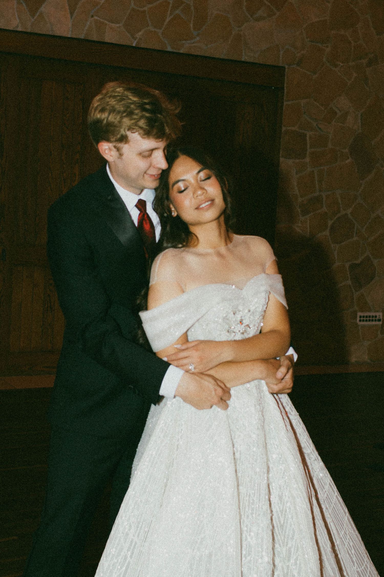 A couple embraces at their wedding, with the bride wearing an elegant off-shoulder white gown and the groom in a dark suit.