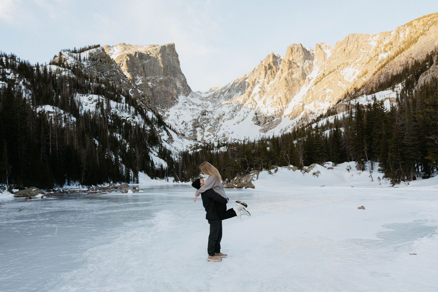 Couple embracing on a frozen lake with snow-capped mountains and evergreen trees in a winter landscape.