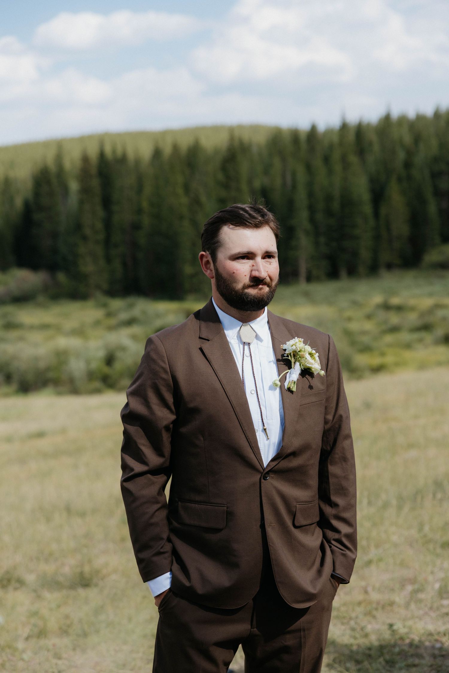 Groom in brown suit with boutonniere stands in mountain meadow with evergreen forest backdrop.