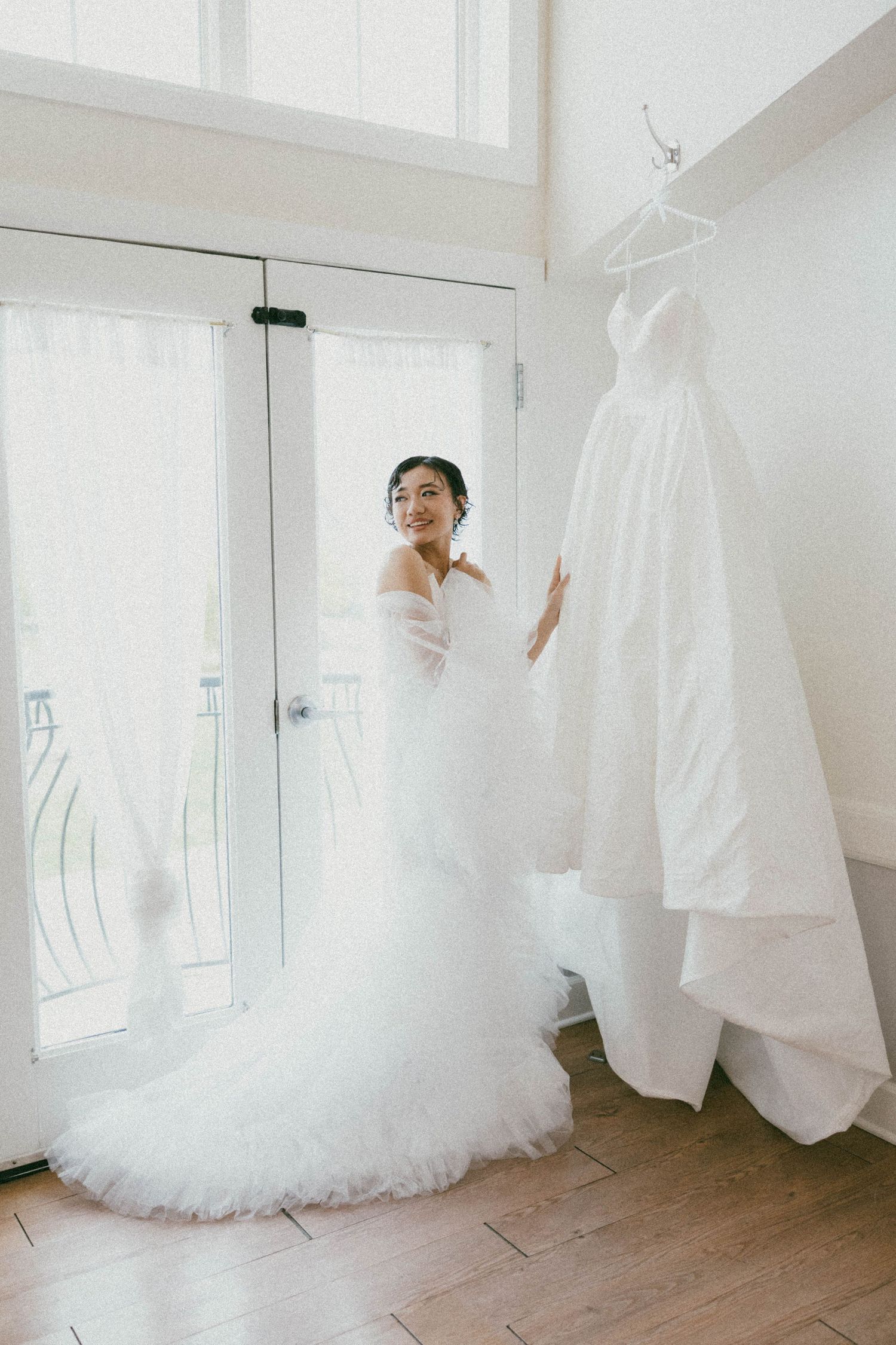 Bride in a white tulle wedding gown standing beside her hanging dress near bright French doors on hardwood floors.