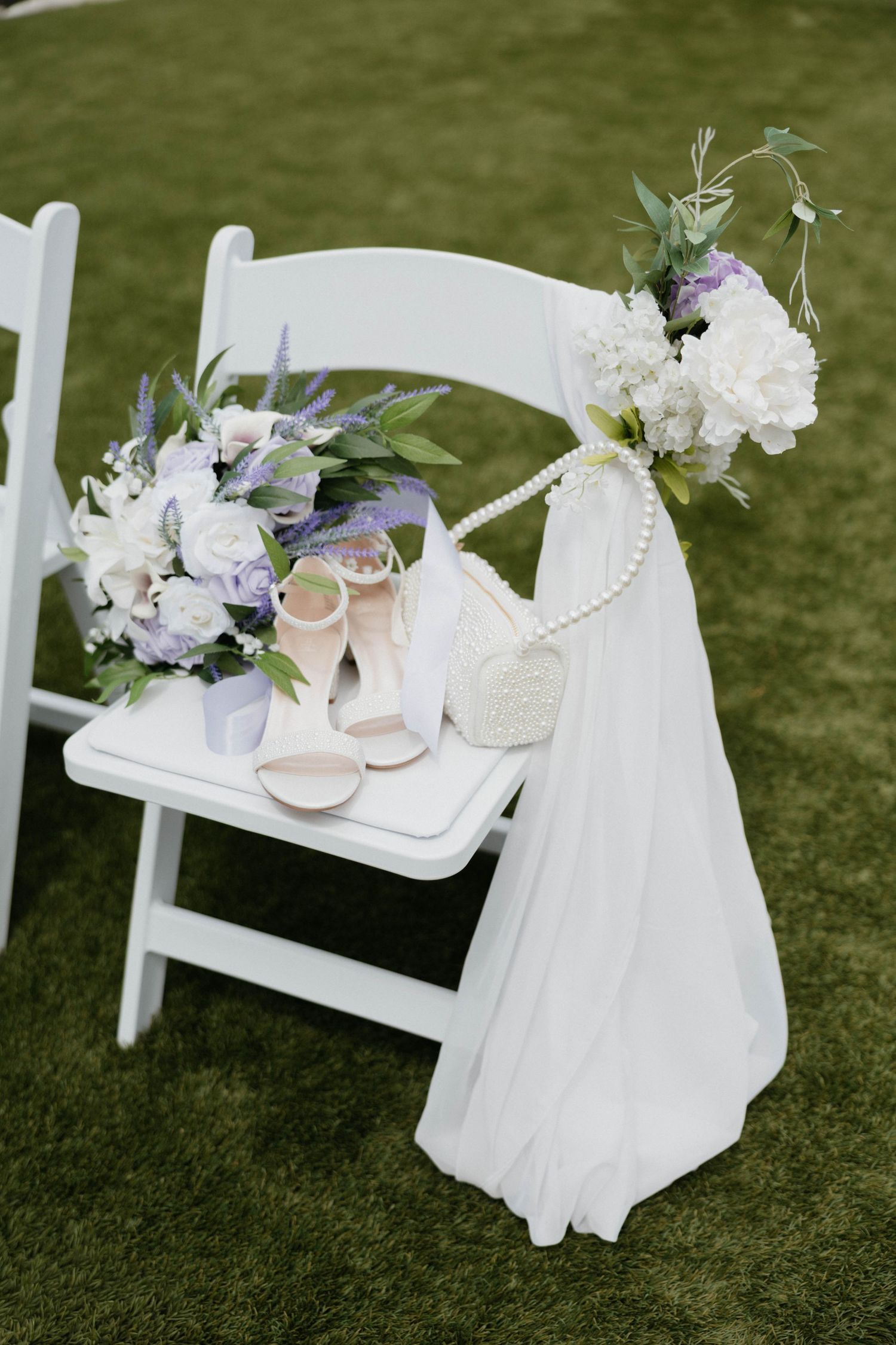 White folding chair with bridal bouquet, nude heels, pearl purse, and white floral arrangement on green grass.