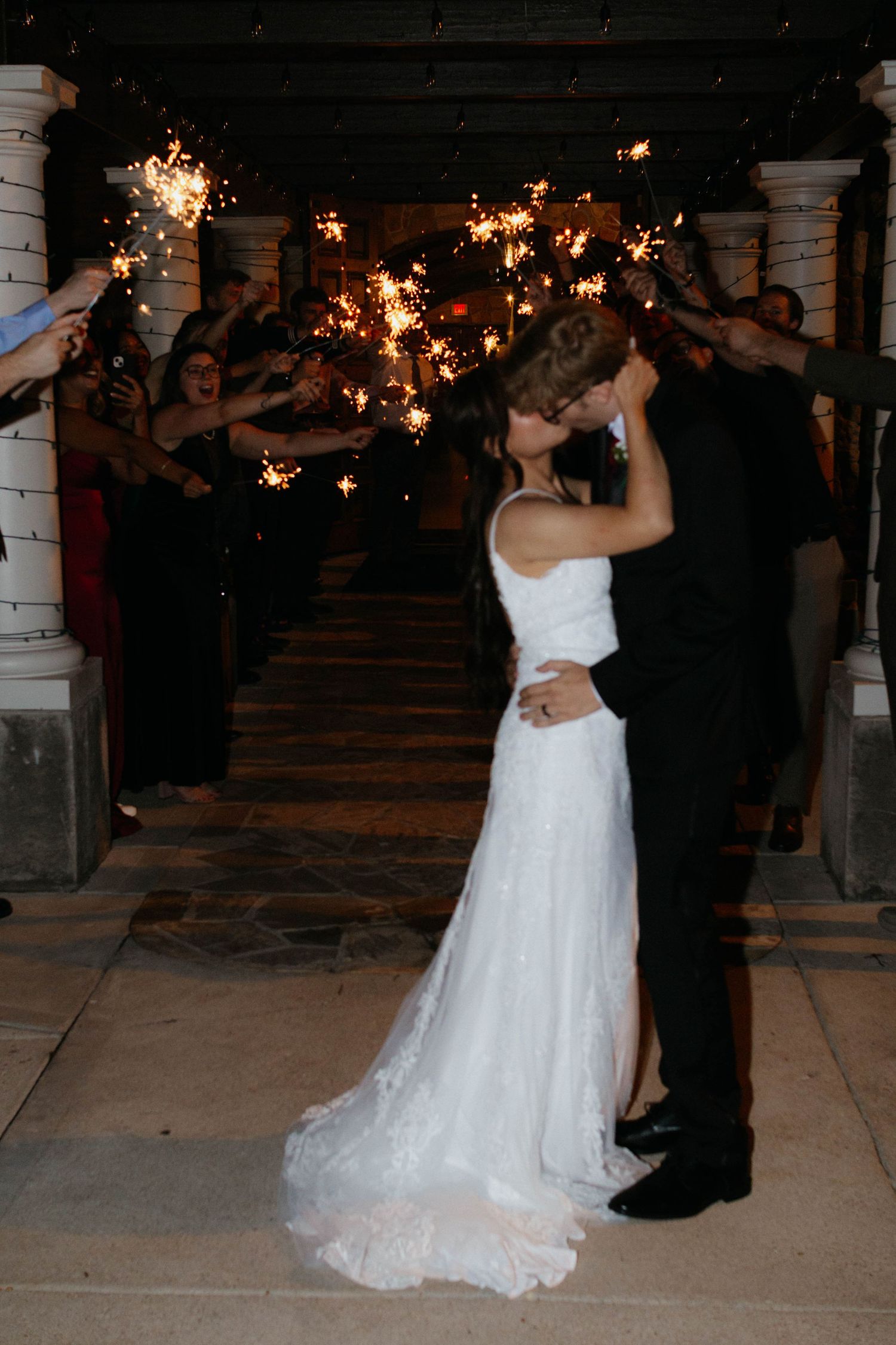 A romantic wedding couple shares a dance under twinkling sparklers at an outdoor venue with white columns.