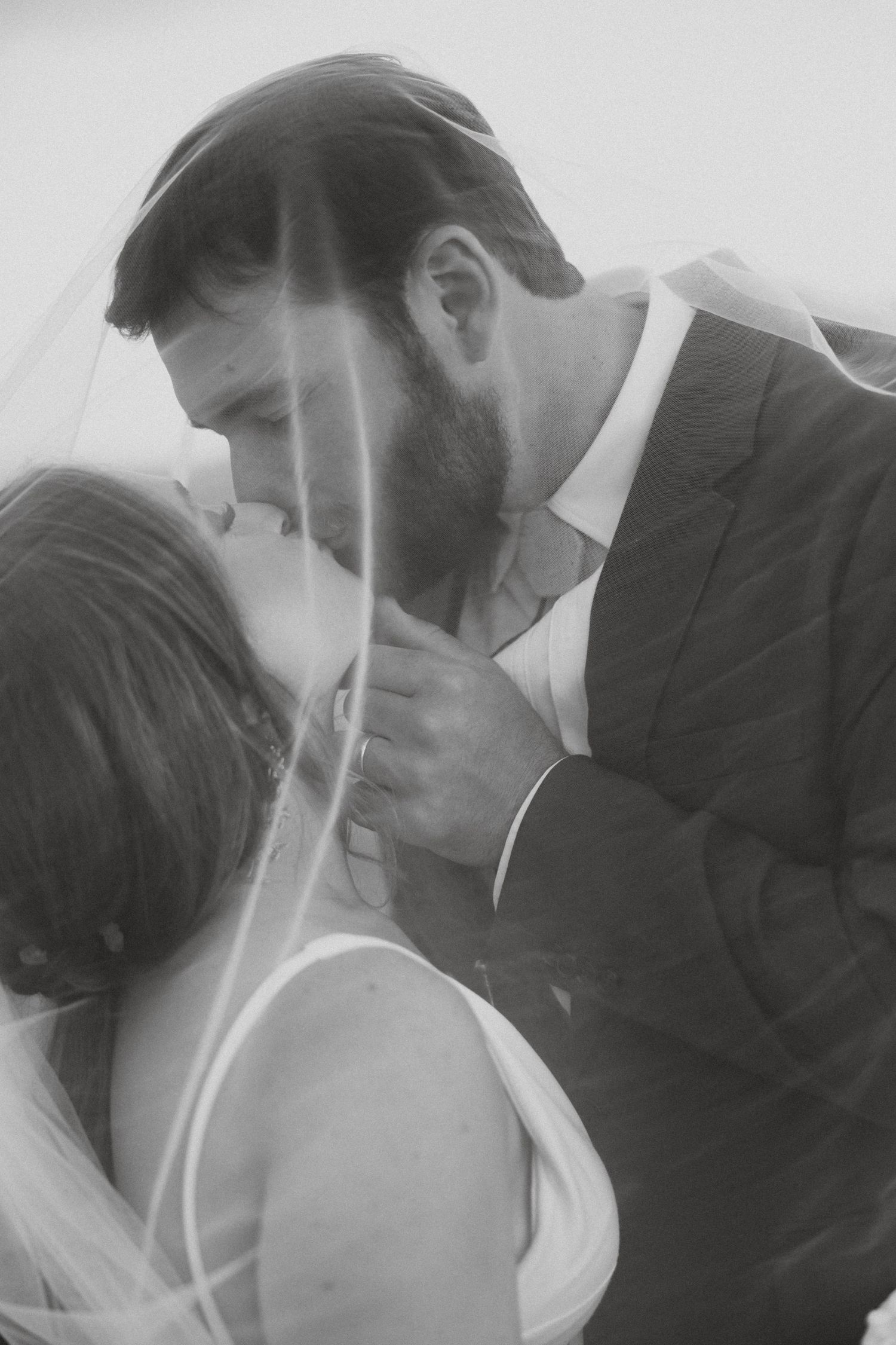 Black and white photo of a bride and groom sharing a romantic kiss under a wedding veil.