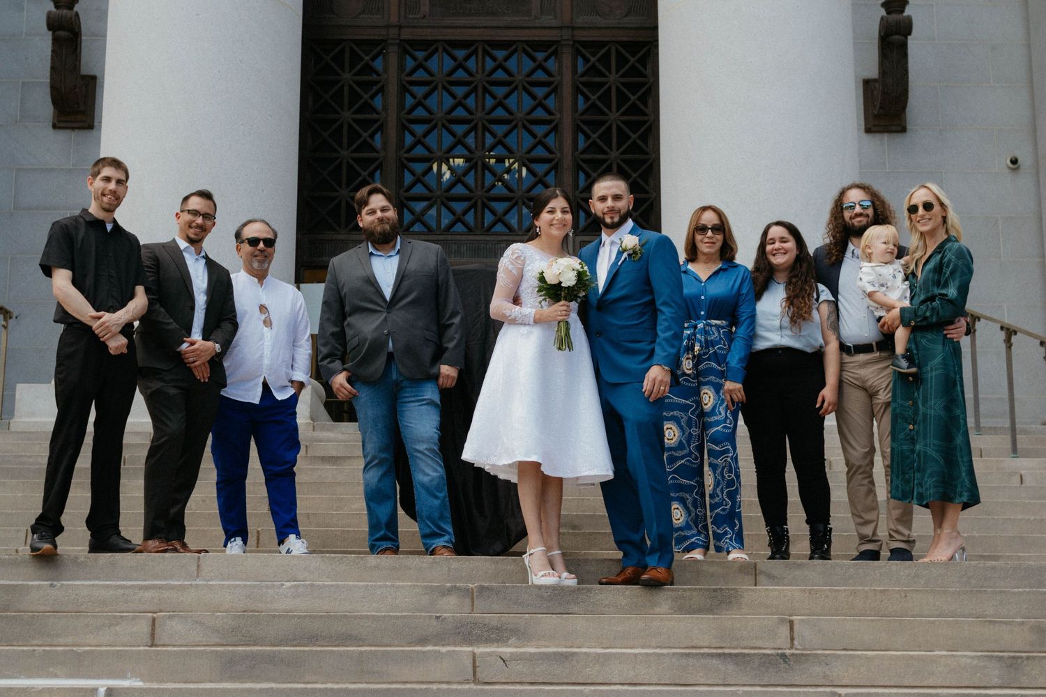 Wedding party poses on steps outside a building, bride in white dress and groom in blue suit surrounded by guests.