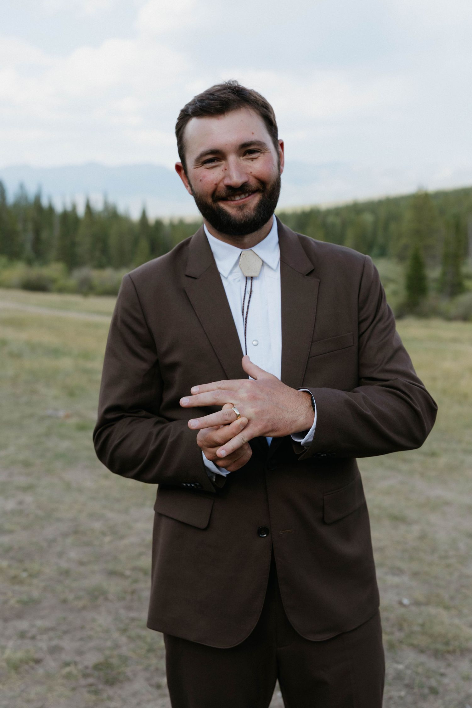 Smiling groom in brown suit with bolo tie clasping hands outdoors with mountain forest backdrop.