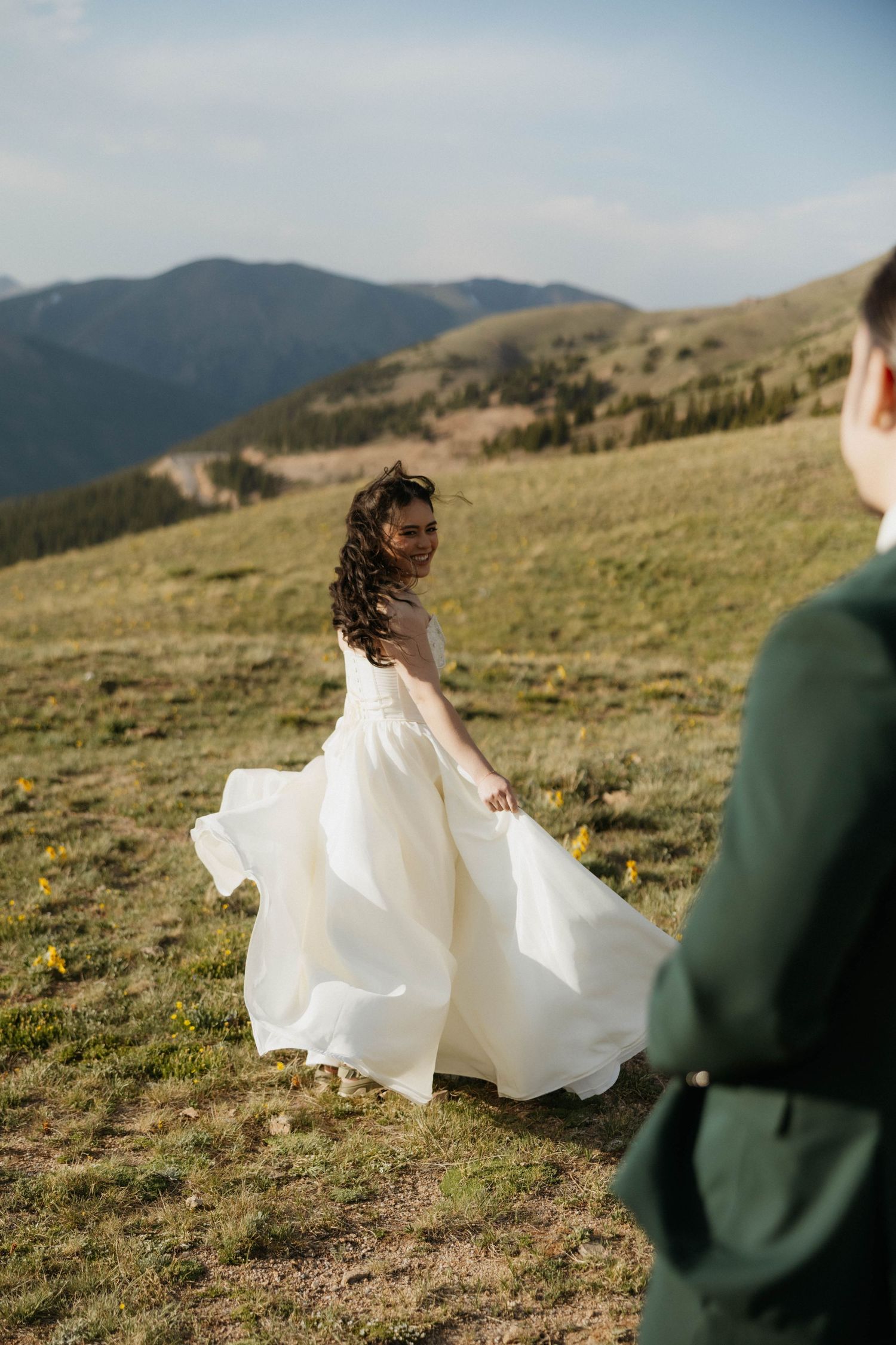 Bride in flowing white gown twirls on mountain meadow, looking back at groom in green suit with scenic hills behind.