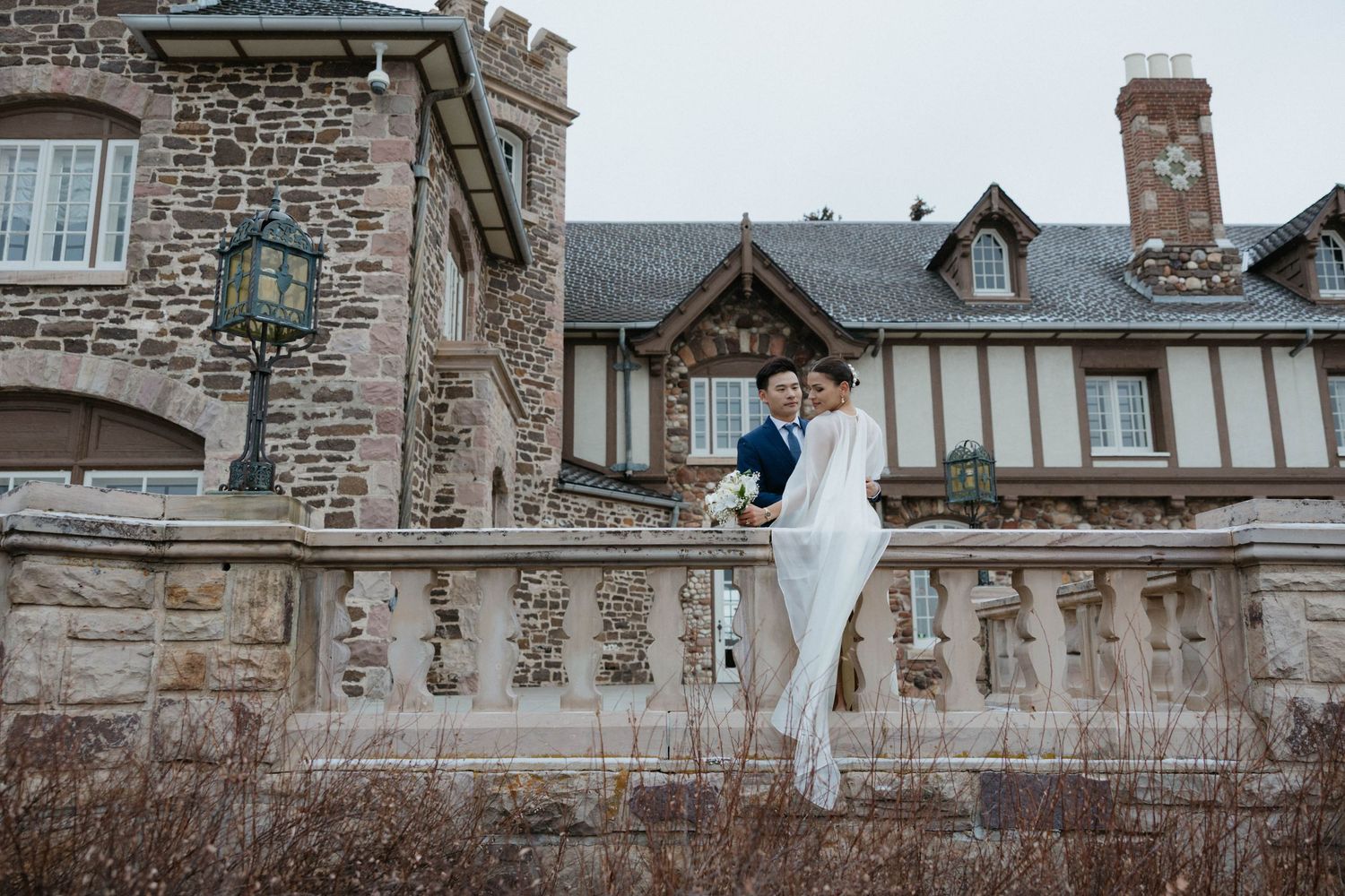 Bride and groom posing elegantly on stone balustrade outside a historic castle venue on their wedding day.
