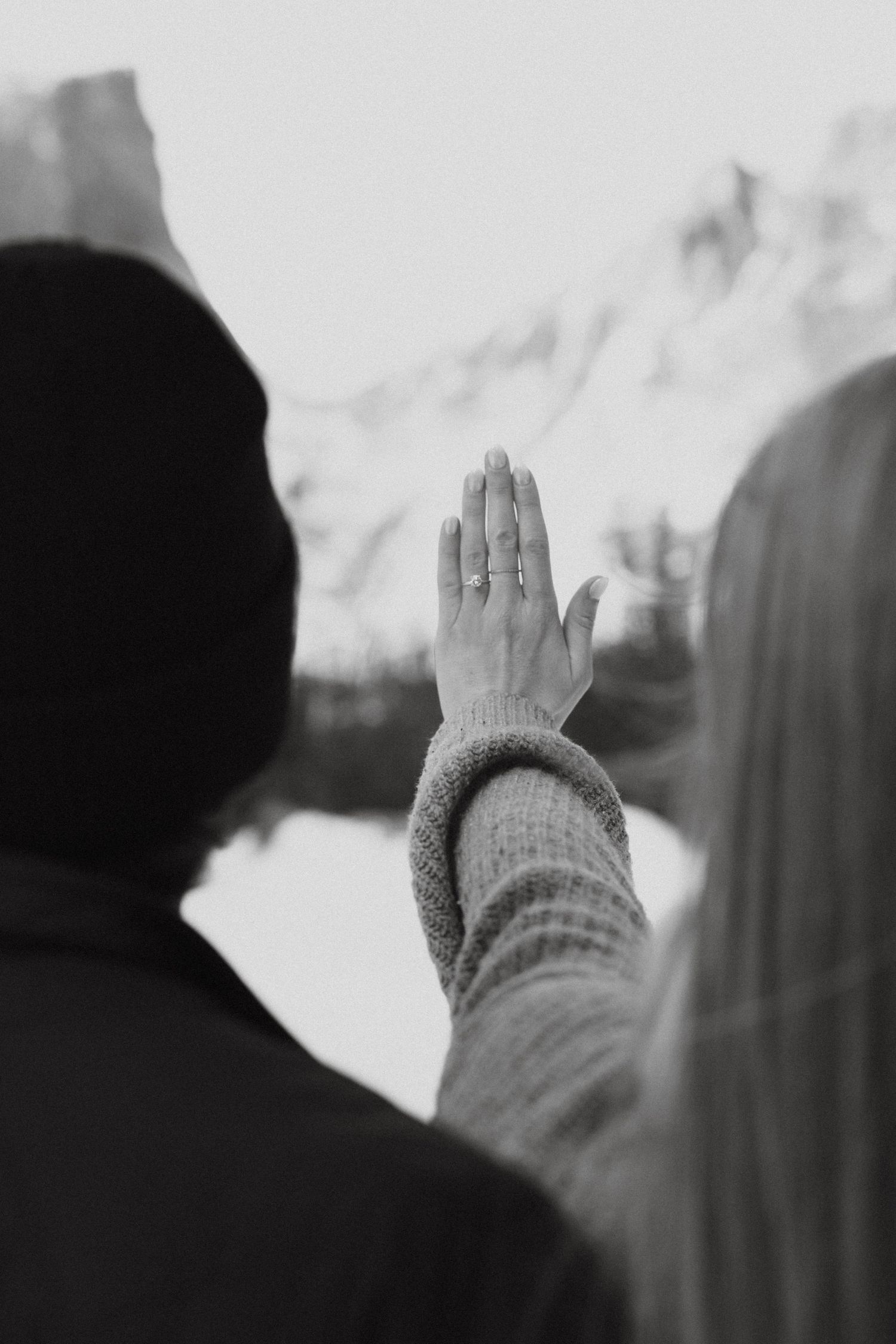 Black and white photo of a woman raising her hand showing an engagement ring with snowy mountains in the background.