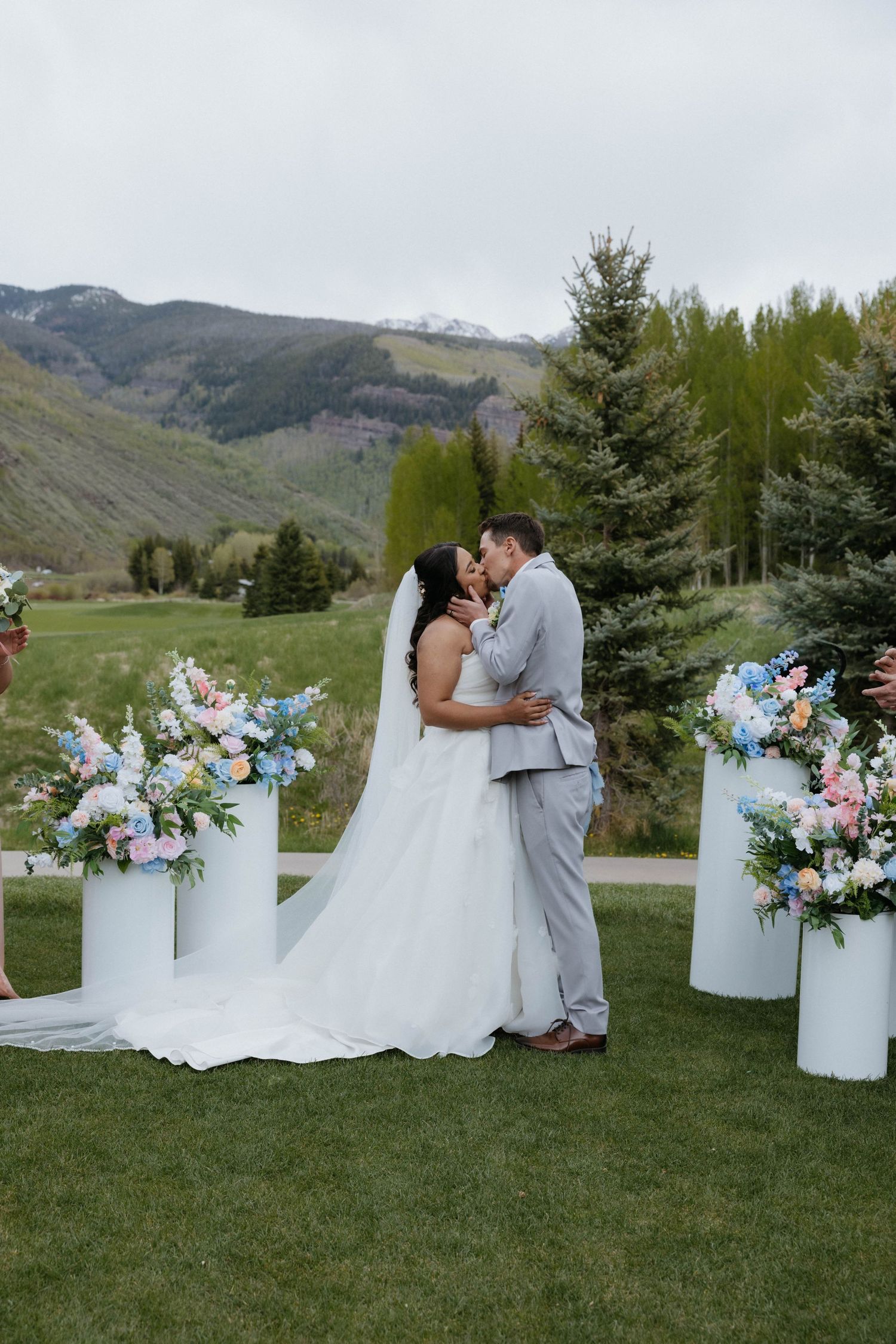 Bride and groom share first kiss at outdoor mountain wedding ceremony with pastel floral arrangements on white pedestals.