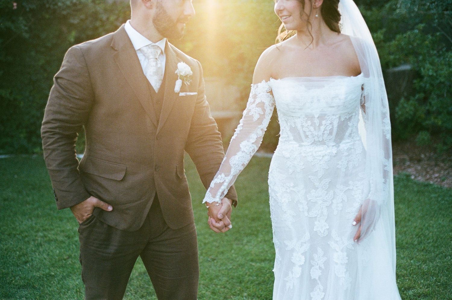 Bride and groom holding hands in golden sunset light, bride wearing lace off-shoulder gown with veil, groom in gray suit.