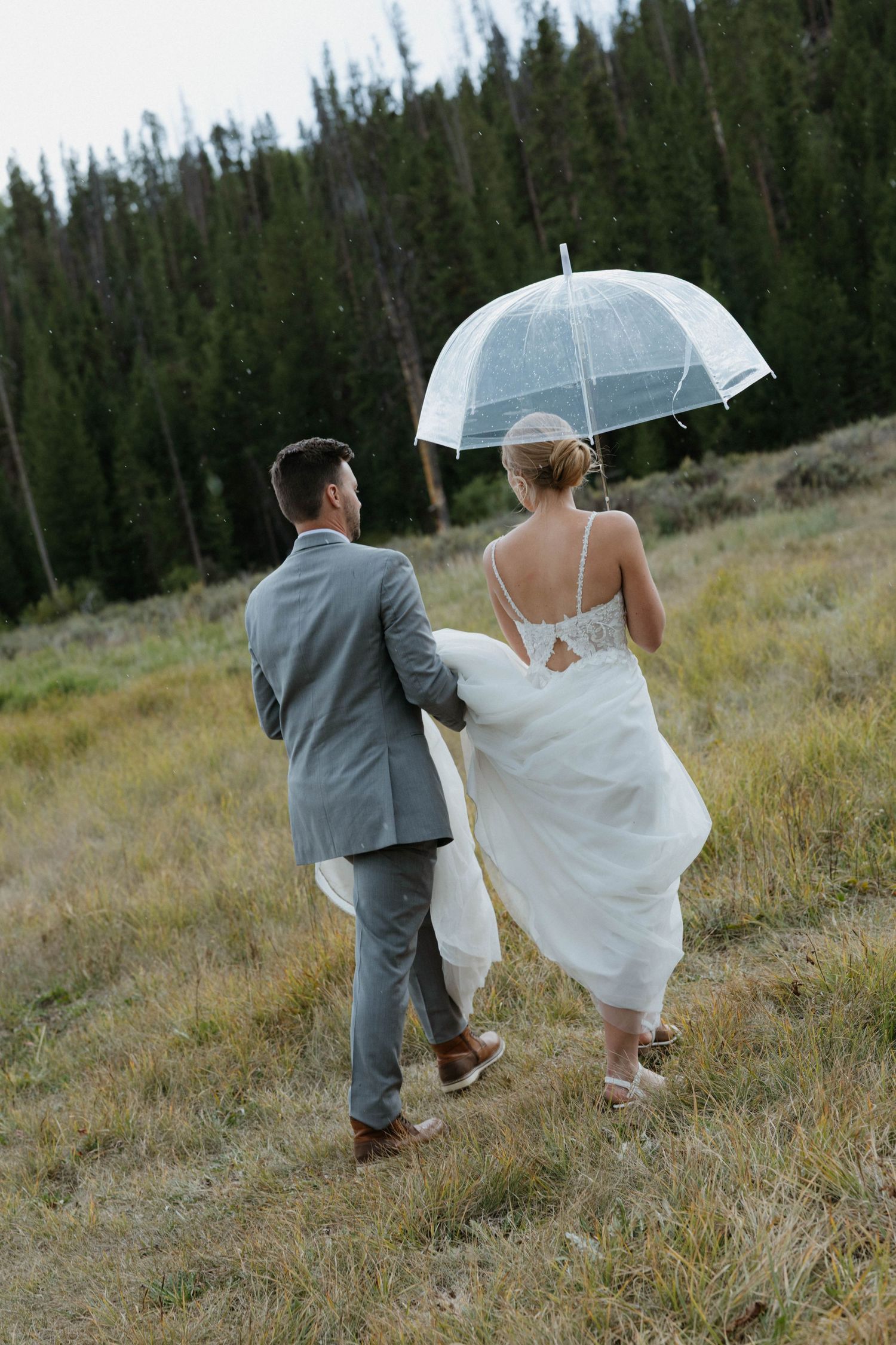 Bride and groom walking through mountain meadow in rain, bride holding clear umbrella wearing backless gown, groom in gray suit.