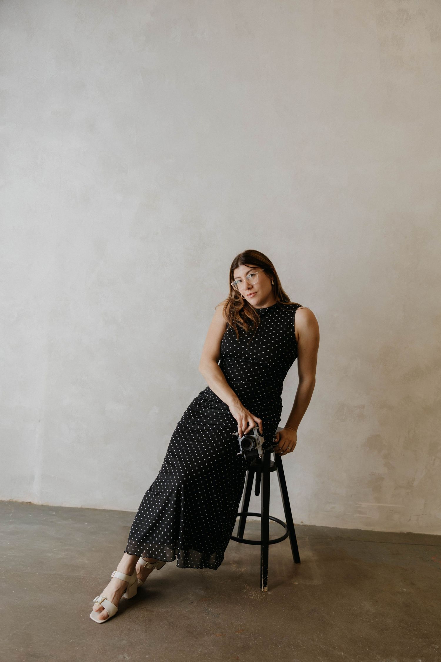 Woman in black polka dot maxi dress seated on a stool, wearing nude sandals against a gray wall.